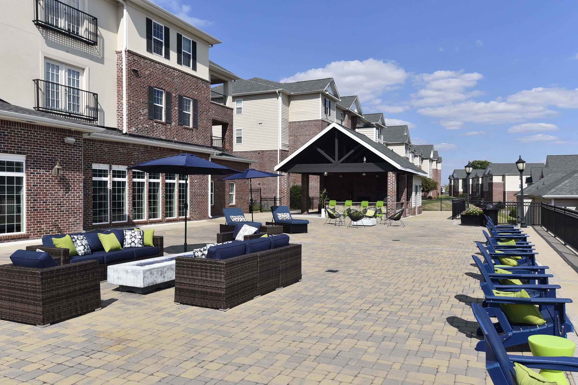 A spacious outdoor patio area featuring modern furniture, including couches with green and blue cushions, and lounge chairs. There's an umbrella for shade and a view of surrounding buildings with a clear blue sky above. The setting appears inviting for relaxation or social gatherings.