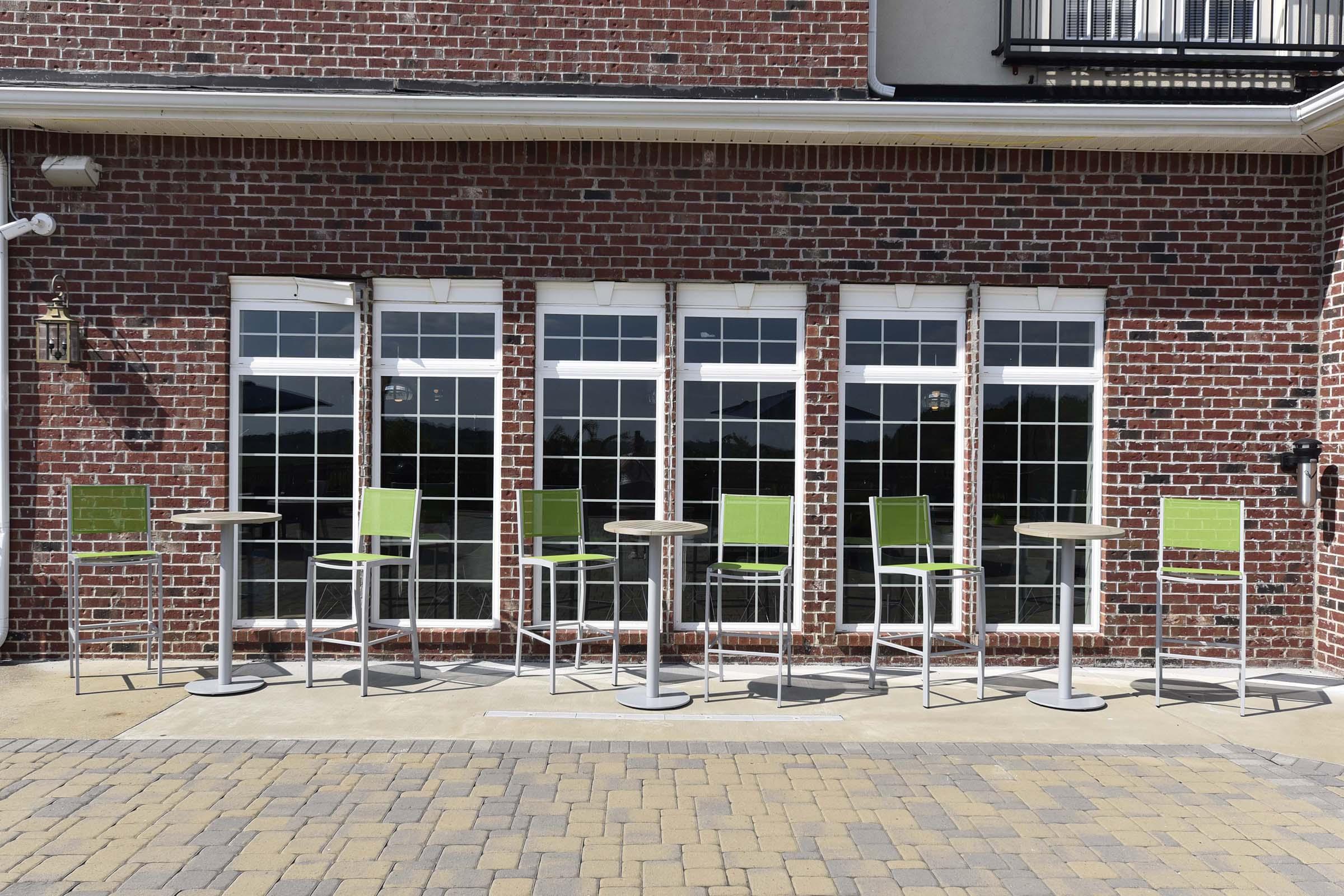 A row of modern green bar stools and high tables set against a brick wall with large windows. The outdoor area features a paved surface with ample natural light, suggesting a welcoming space for relaxation or socializing.