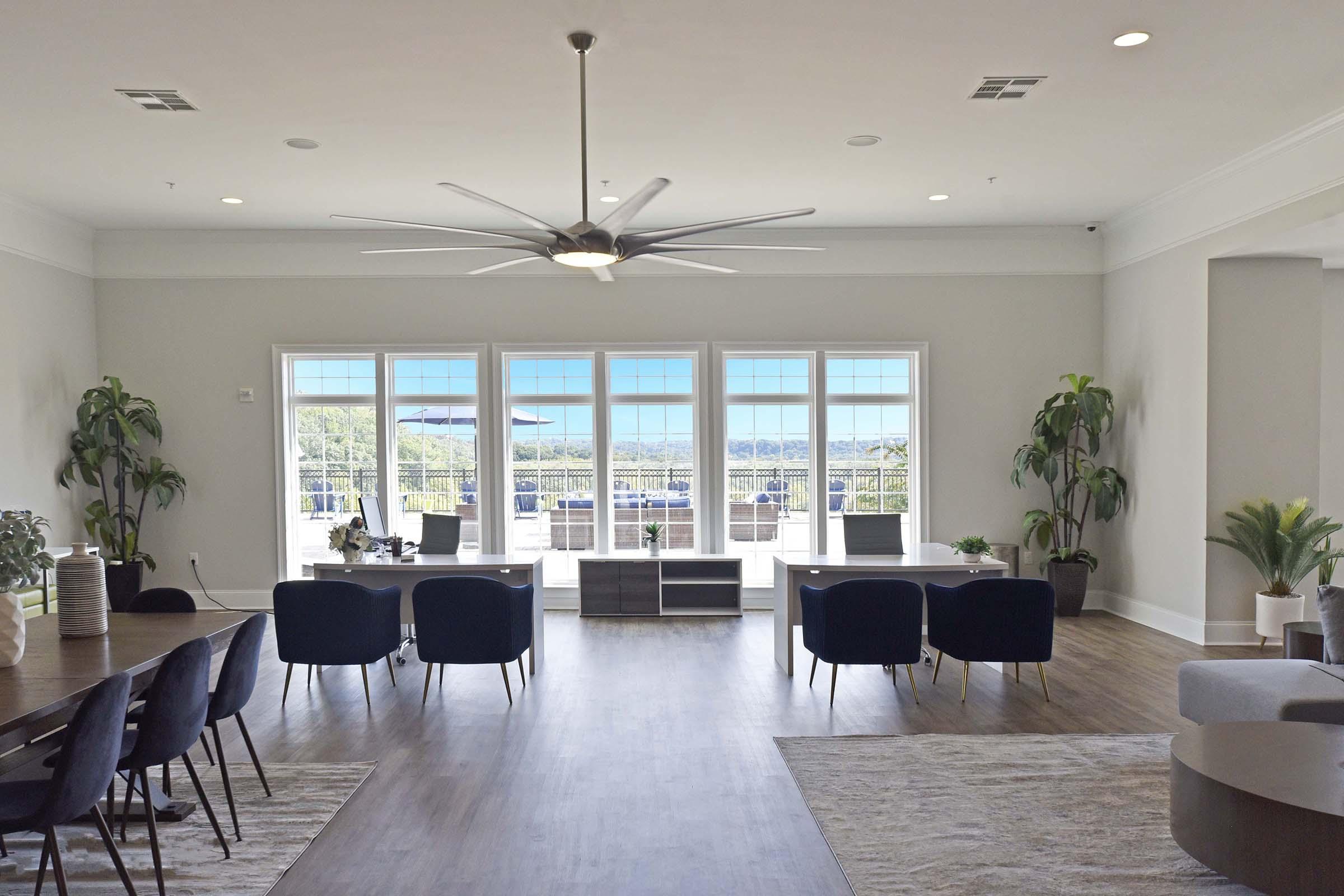 A modern living space featuring a large open area with a wooden floor, several indoor plants, and comfortable seating. Large windows offer a view of the outdoors, while a dining table with chairs and a sleek entertainment unit adds to the contemporary design. Natural light brightens the room.