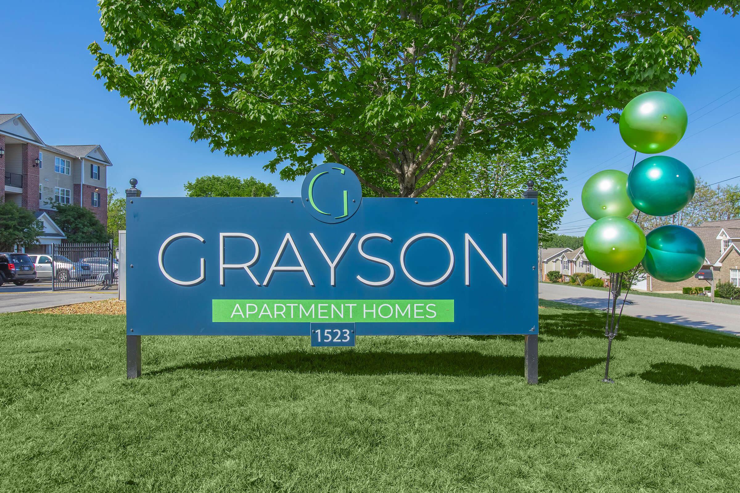 A blue and green sign displaying "GRAYSON APARTMENT HOMES" with the number "1523" at the bottom. The sign is surrounded by lush green grass and features a decorative arrangement of green and blue balloons nearby, with a tree visible in the background against a clear sky.