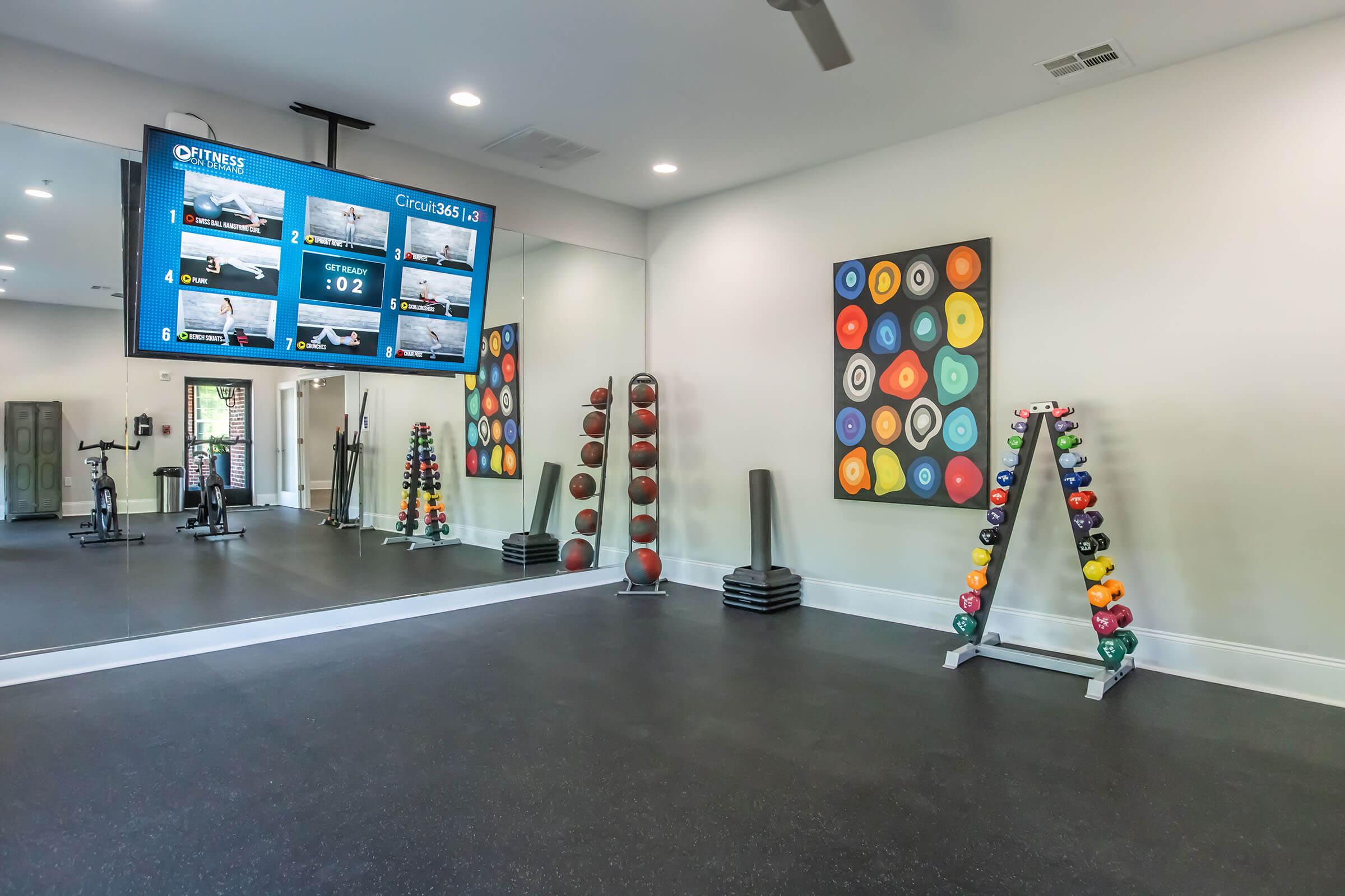 A modern fitness room featuring a large wall-mounted screen displaying workout stats, a mirrored wall, and various exercise equipment. On one side, there are colorful weight balls arranged in a rack, while the opposite side has other fitness equipment, creating an inviting workout space.