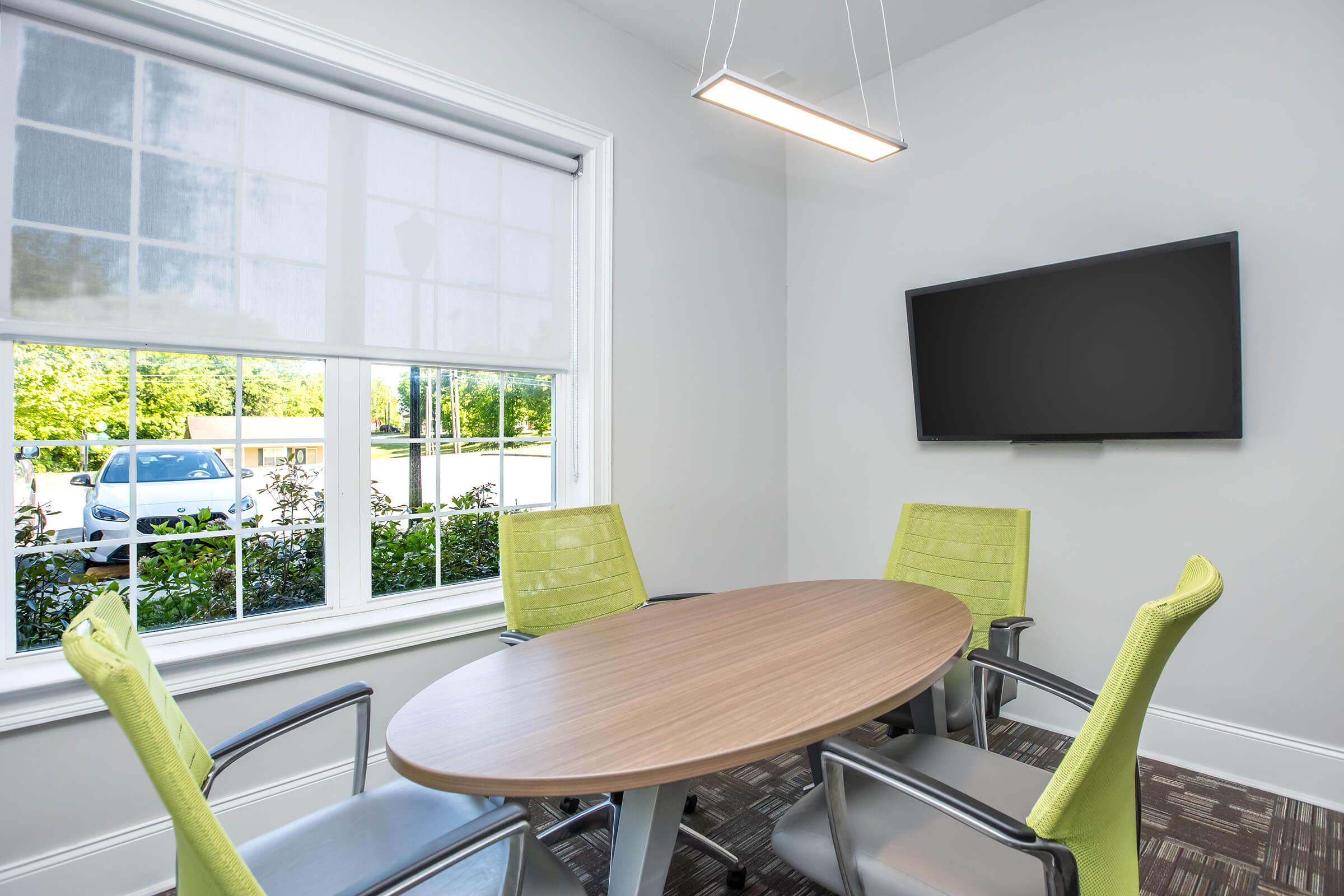 A modern conference room featuring a round wooden table surrounded by four green office chairs. A large wall-mounted TV is visible, and natural light streams in from a window with shades, overlooking a landscaped area with greenery.