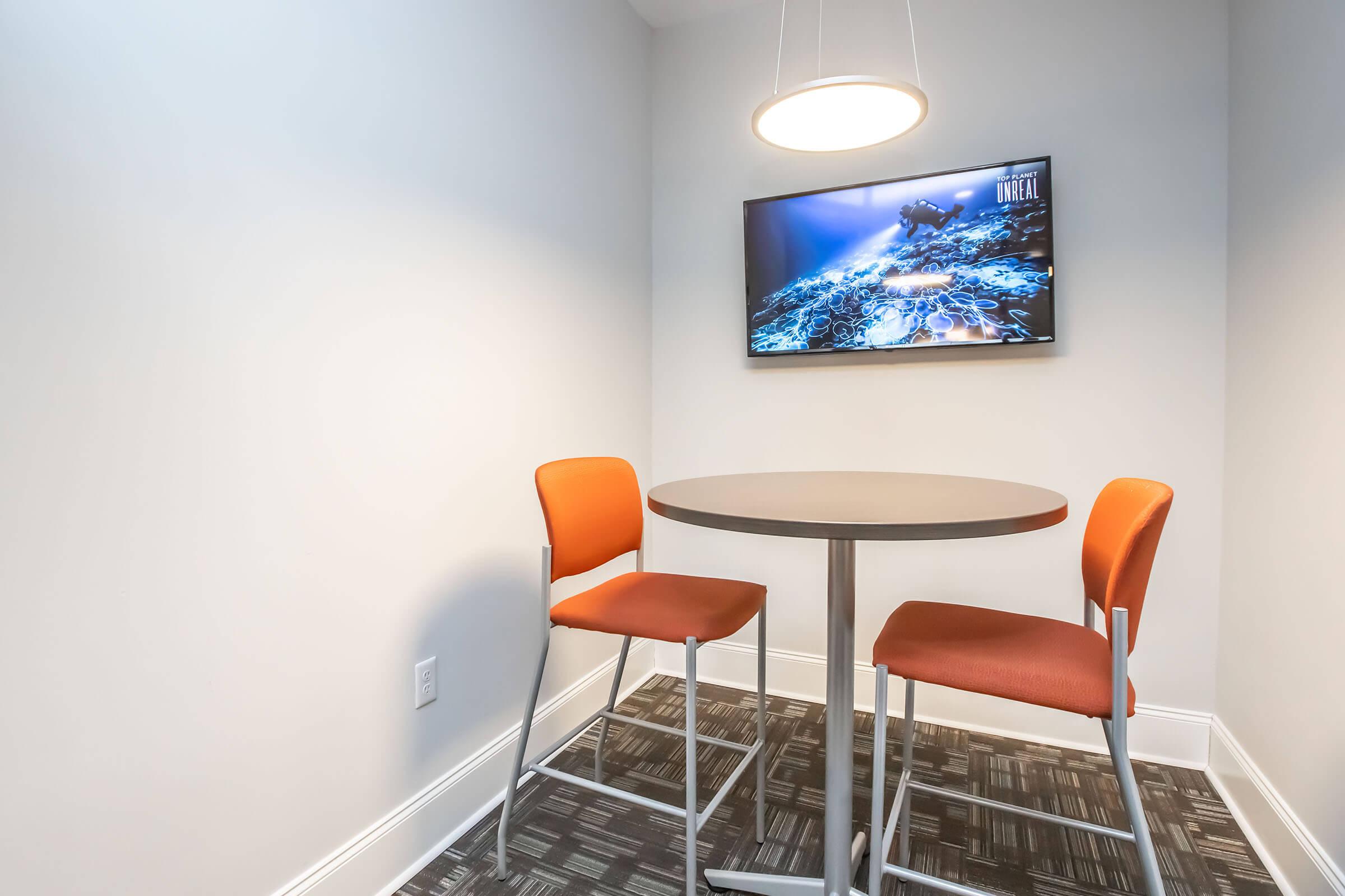 A small meeting space featuring a round table with two orange chairs. A flat-screen TV mounted on the wall displays an underwater scene. The room has neutral-colored walls and a patterned floor, illuminated by a modern ceiling light.
