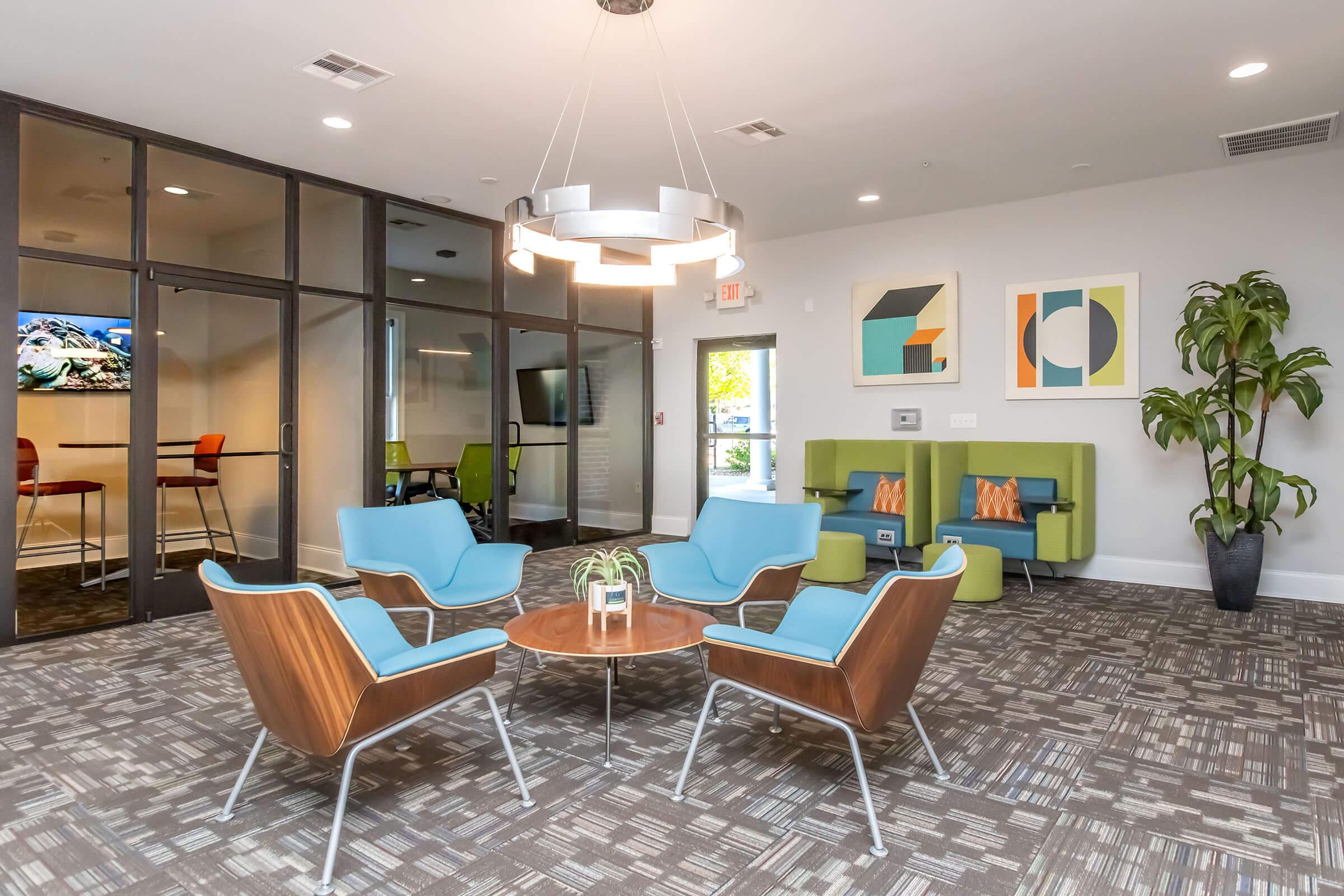 A modern lounge area featuring four blue chairs arranged around a round table. Nearby, there is a green couch with decorative pillows, and colorful wall art is displayed. A large window offers natural light, and there are plants adding greenery to the space. Glass doors lead to a conference room visible in the background.