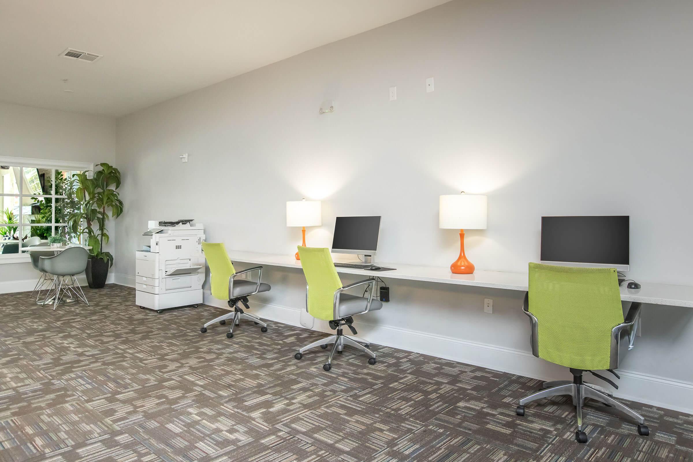 A modern workspace featuring three desks with computers and ergonomic chairs. Each desk has a stylish lamp with an orange base. A printer is located nearby, and there is a large window with natural light and a potted plant, creating a bright and inviting atmosphere. The flooring has a patterned carpet.