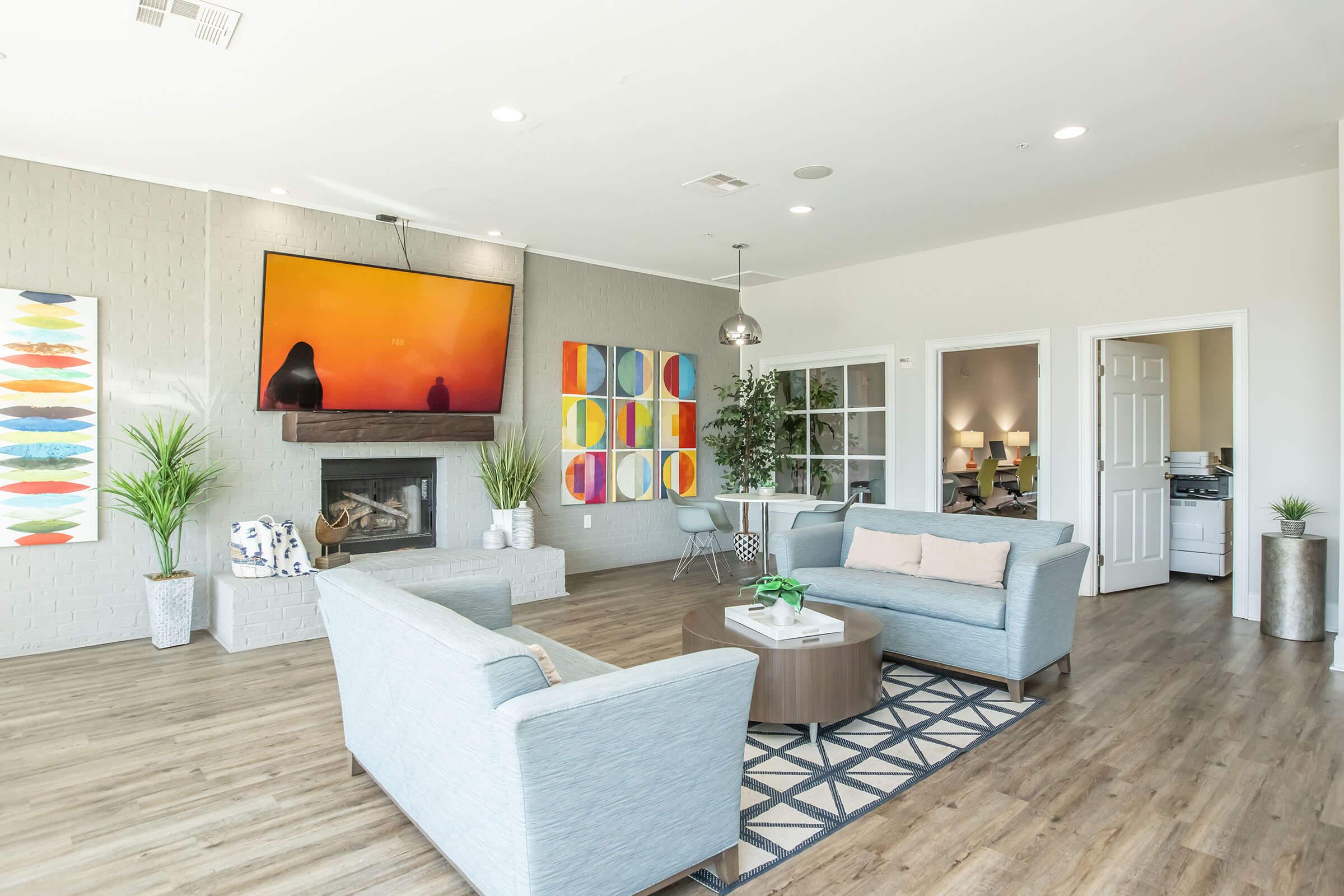 A modern living room featuring light blue sofas and a coffee table on a geometric rug. There is a flat-screen TV mounted above a fireplace, with vibrant abstract artwork on the walls. Potted plants add a touch of greenery, and a glass door leads to another room, creating an inviting and stylish atmosphere.