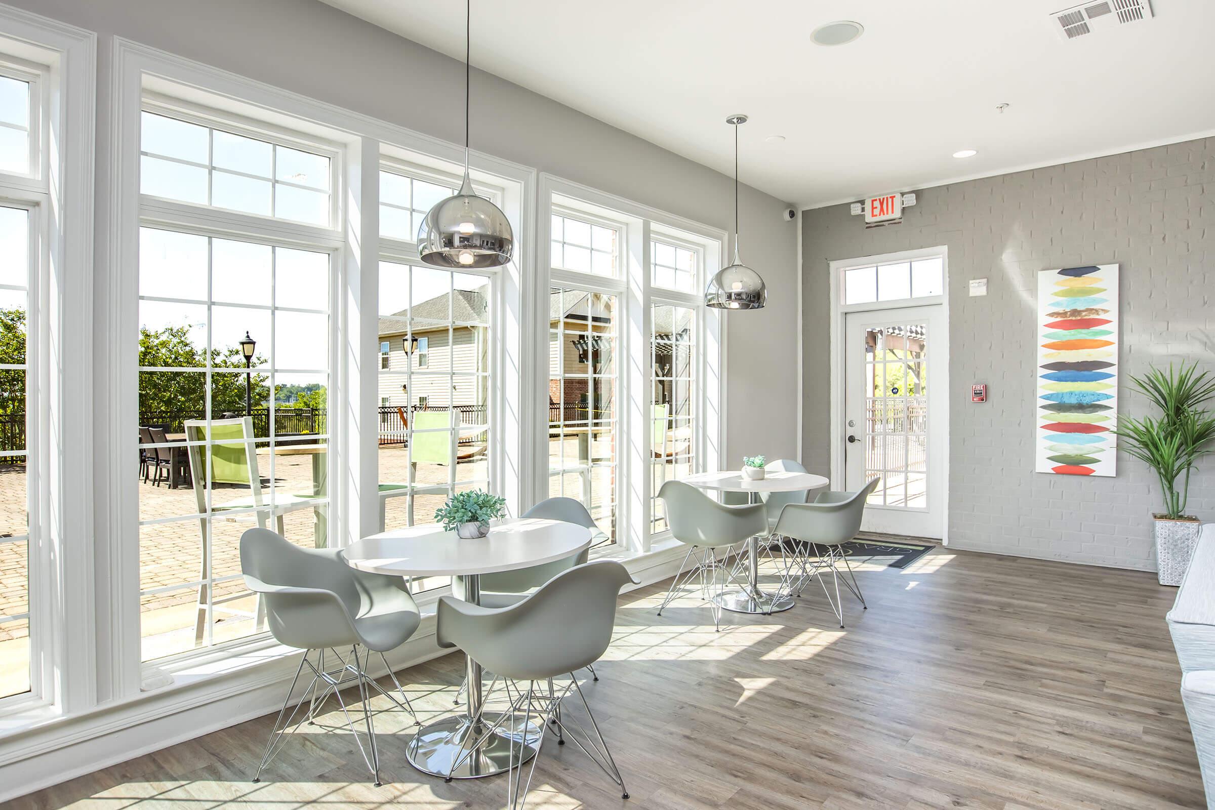 Bright, modern interior of a café with large windows allowing natural light. The space features round white tables with grey chairs, hanging metallic light fixtures, and a decorative wall with colorful art. Potted plants add a touch of greenery, while the scenic outdoor view can be seen through the windows.