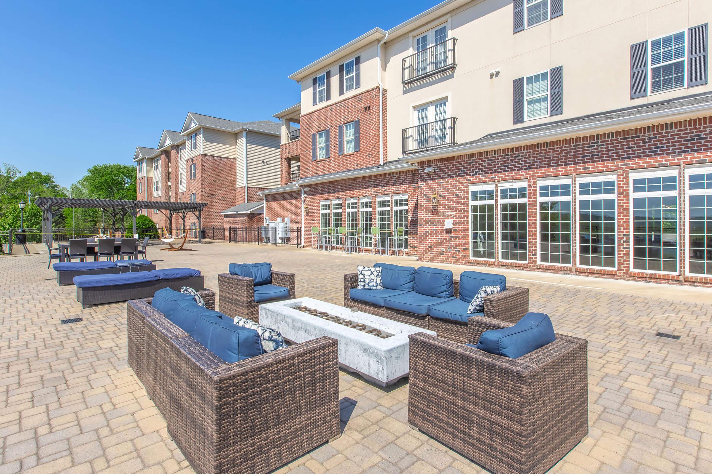 A spacious outdoor patio area featuring comfortable seating with blue cushions, a central fire pit, and an inviting atmosphere. The background includes a multi-story building with large windows and a pergola, set against a clear blue sky and greenery. Perfect for relaxation and social gatherings.