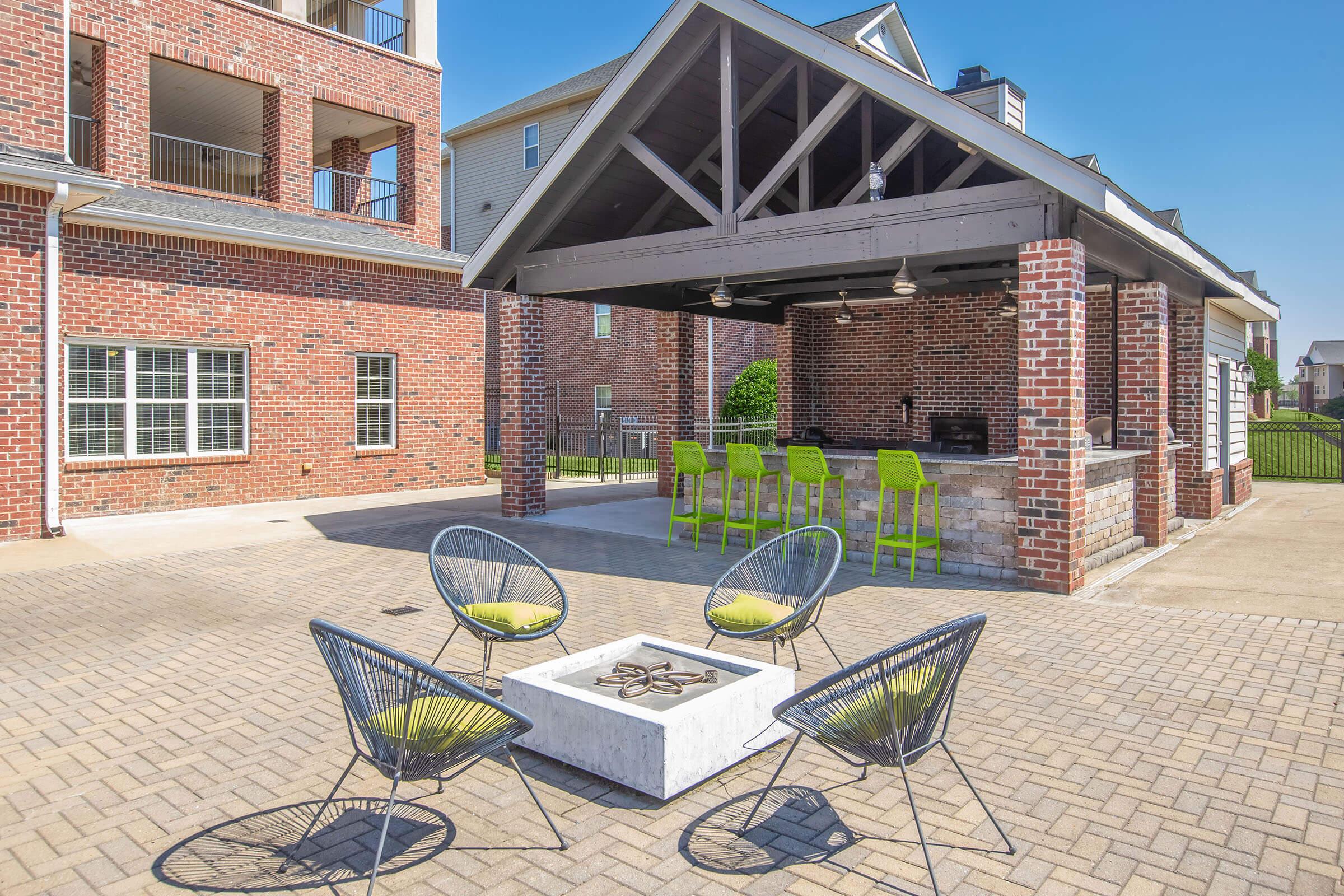 An outdoor communal area featuring a covered pavilion with bar seating, surrounded by brick buildings. In the foreground, there is a circular fire pit with modern chairs, all situated on a patterned stone patio. The scene is bright and sunny, creating a welcoming atmosphere.