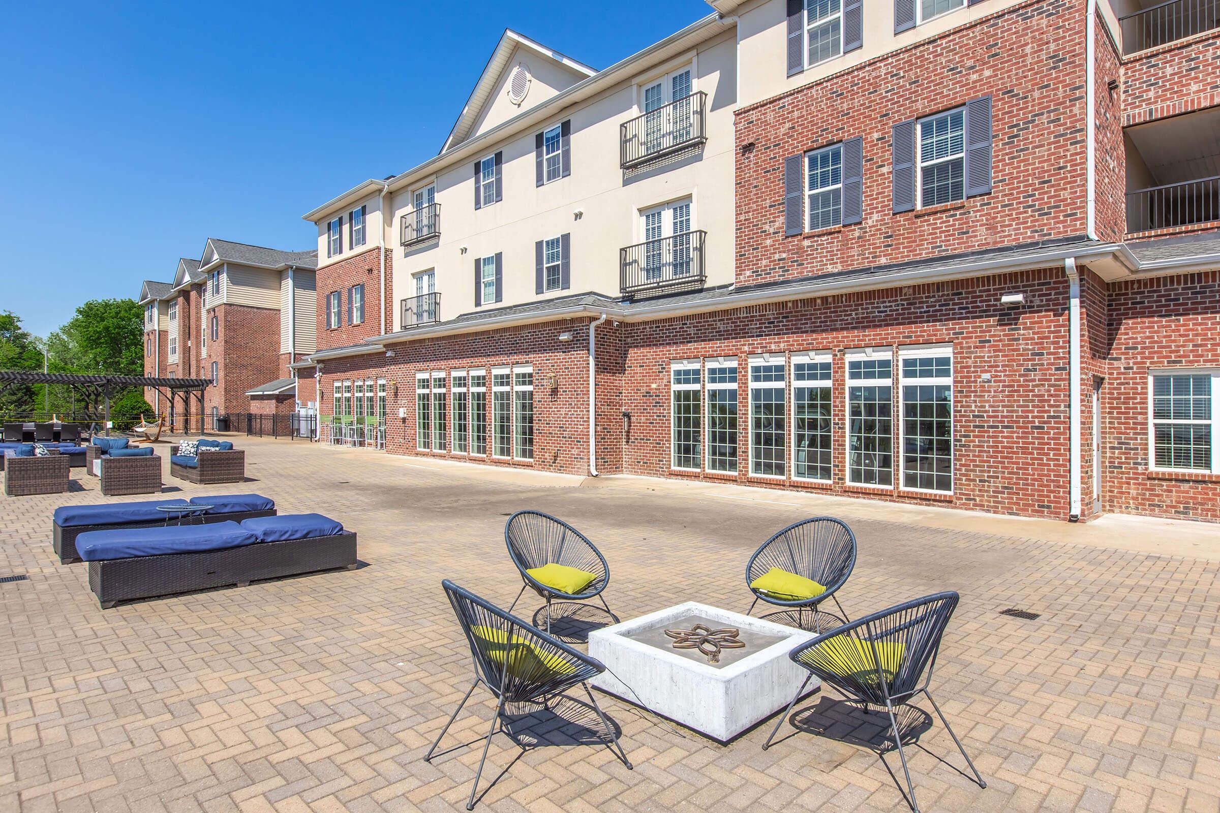 An outdoor patio area featuring modern seating with round chairs and a fire pit, surrounded by apartment buildings with brick facades. The clear blue sky and well-maintained landscaping enhance the inviting atmosphere of the space.