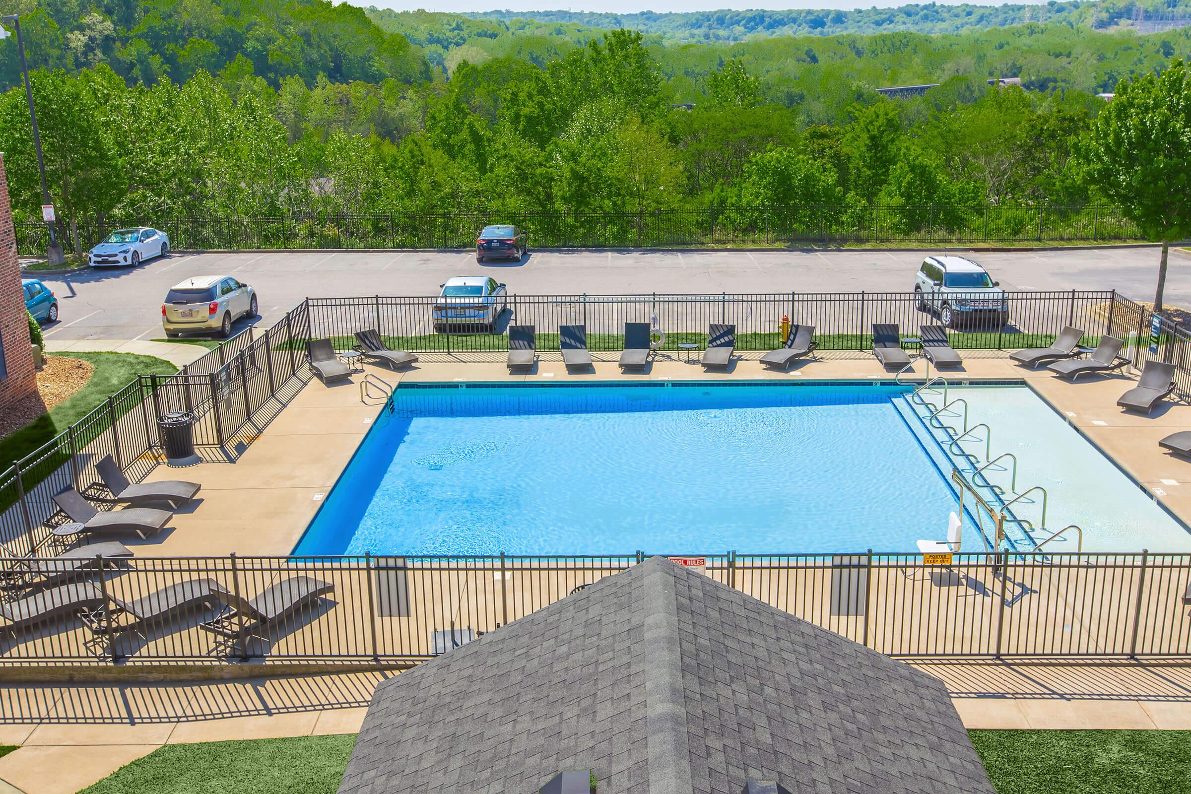 A clear outdoor swimming pool surrounded by lounge chairs and a fence, with lush green trees in the background. Several cars are parked nearby, and the scene is bright and sunny, suggesting a pleasant recreational area.