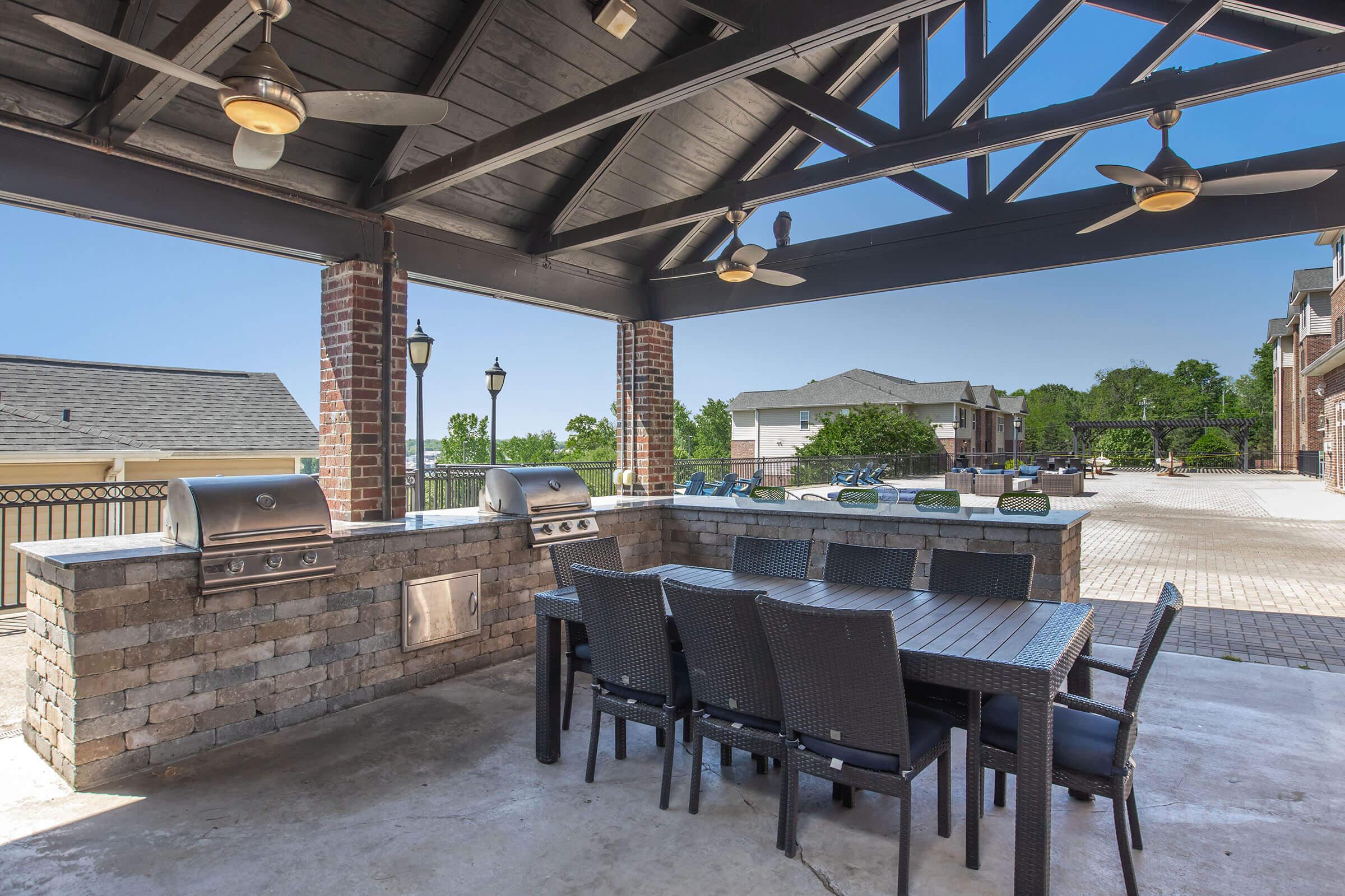 Outdoor kitchen with a covered seating area featuring a dining table and chairs. Equipped with multiple stainless steel grills and a stone countertop. Bright blue sky and trees visible in the background, along with a patio area and nearby residential buildings.