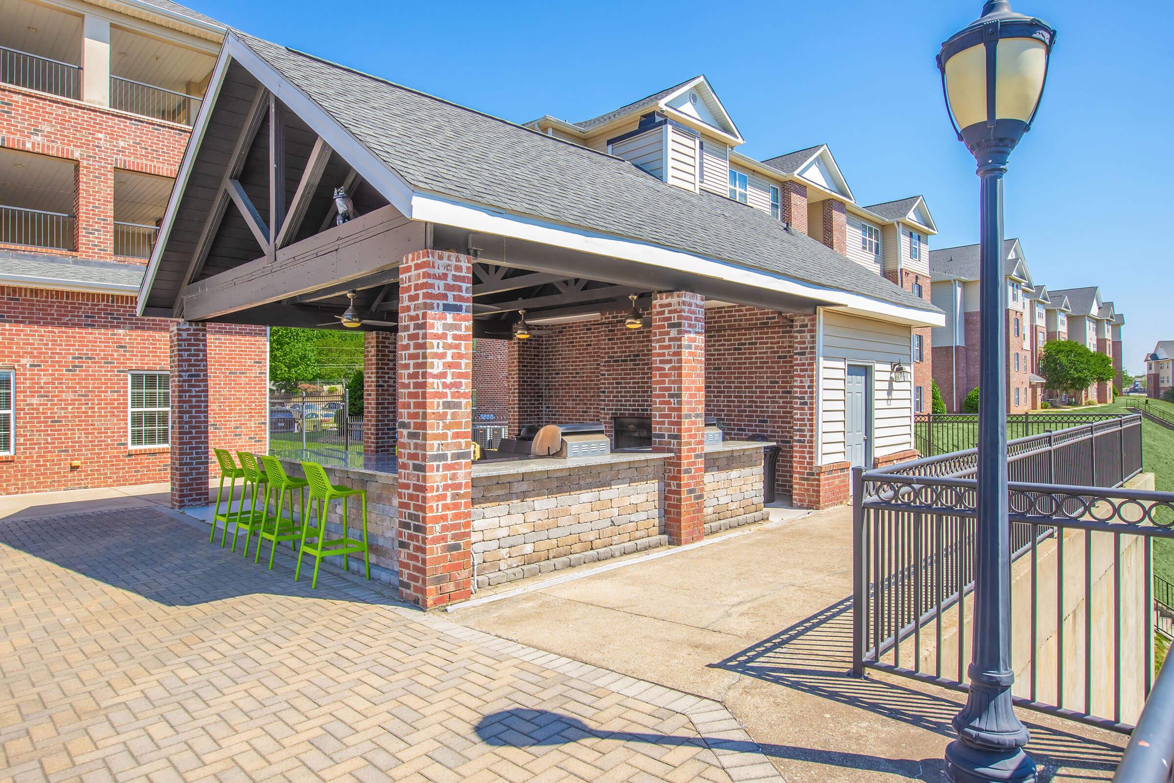 A modern outdoor pavilion with a gray roof and brick walls features a built-in barbecue area and seating. Bright green chairs are arranged around a stone bar. The surrounding area includes a landscaped lawn and residential buildings under a clear blue sky, creating a welcoming atmosphere for outdoor gatherings.