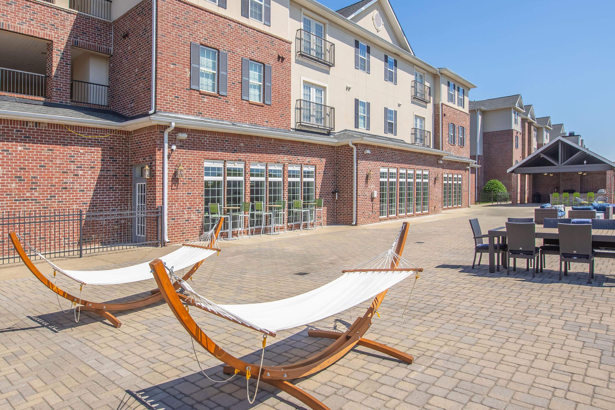 Two hammocks are positioned on a paved patio in front of a brick building with multiple windows and balconies. In the background, there are outdoor seating areas and a shaded pavilion. The sky is clear, indicating a sunny day.