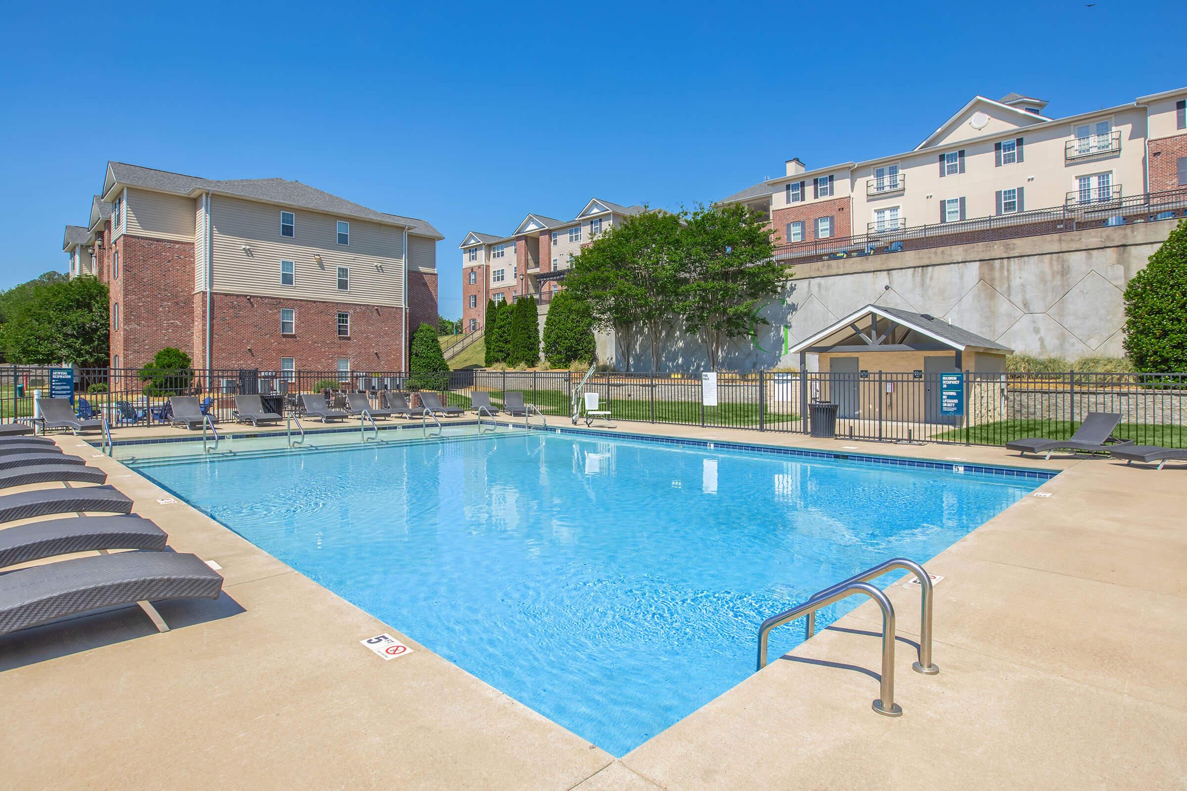 A clear blue swimming pool surrounded by lounge chairs, with brick apartment buildings in the background. The sky is bright and sunny, enhancing the outdoor relaxation space. The pool area features a paved deck and a fence for safety.