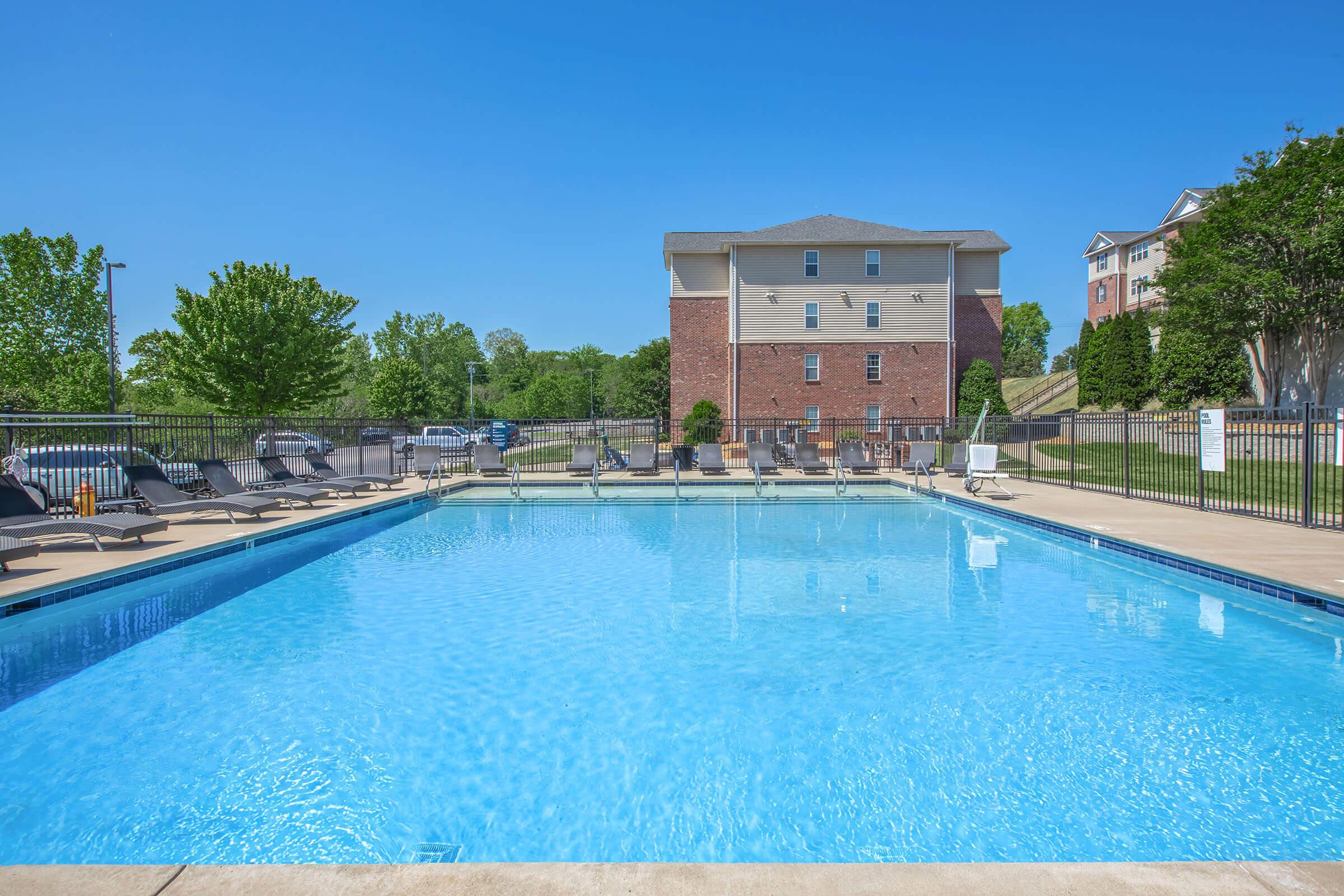 A clear blue swimming pool surrounded by lounge chairs, with a grassy area and trees in the background. A multi-story brick building is visible behind the pool, under a bright blue sky. The scene conveys a relaxing outdoor atmosphere.