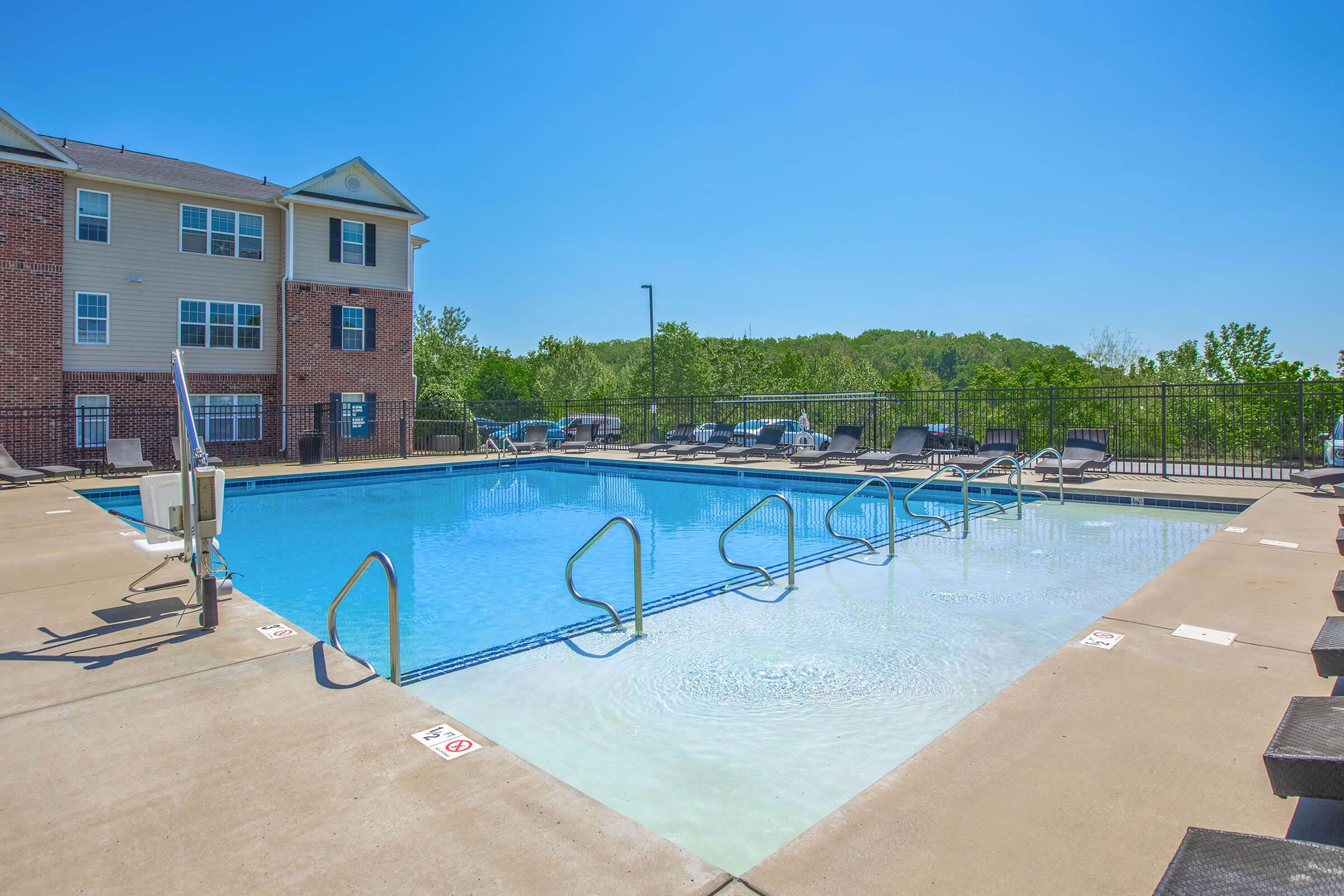 A clear swimming pool surrounded by a concrete deck, with lounge chairs arranged along the edge. In the background, a multi-level apartment building with several windows is partially visible. The scene is set on a sunny day with a blue sky and greenery in the distance.