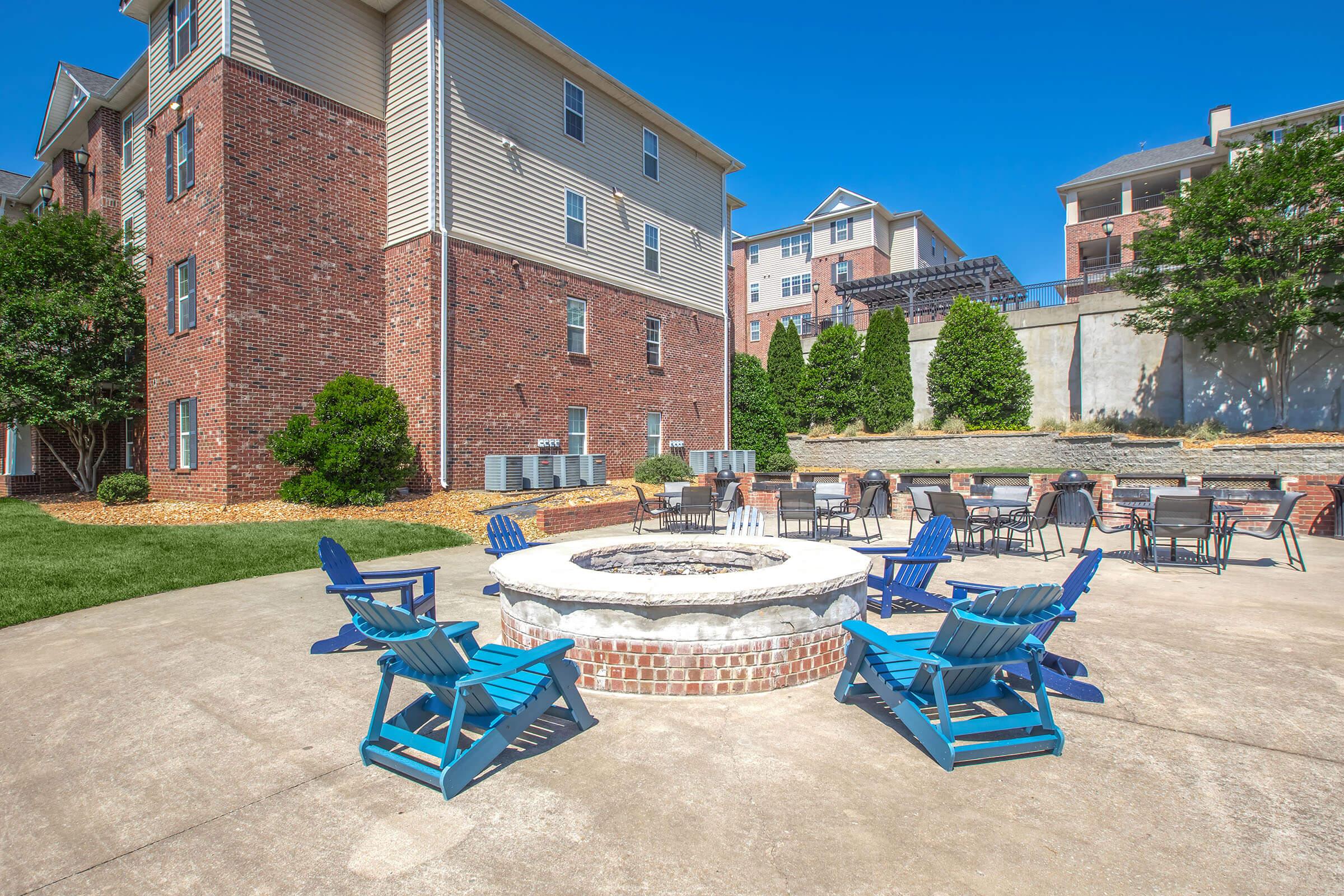 A circular brick fire pit surrounded by blue Adirondack chairs in an outdoor area. The setting features a neatly landscaped lawn, with brick buildings in the background and a sunny blue sky overhead. Tables and chairs are set nearby, creating a cozy gathering space.