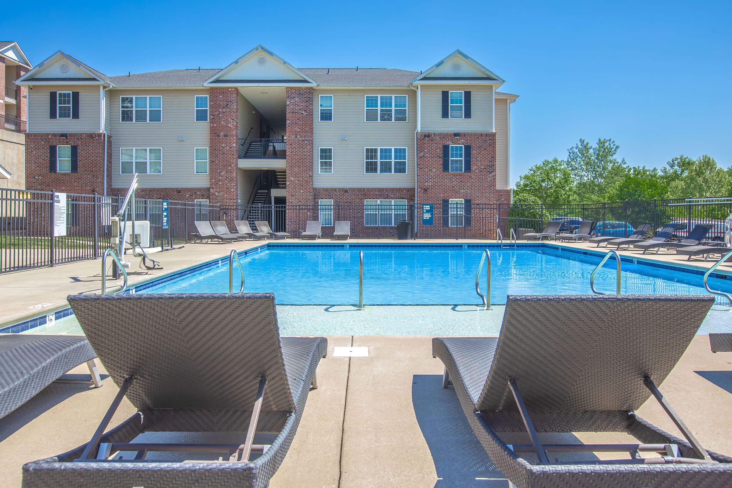 A clear blue swimming pool surrounded by lounge chairs, with a multi-story apartment building in the background. The sky is sunny and bright, enhancing the inviting atmosphere of the outdoor space. A fence encircles the pool area, providing a private retreat.