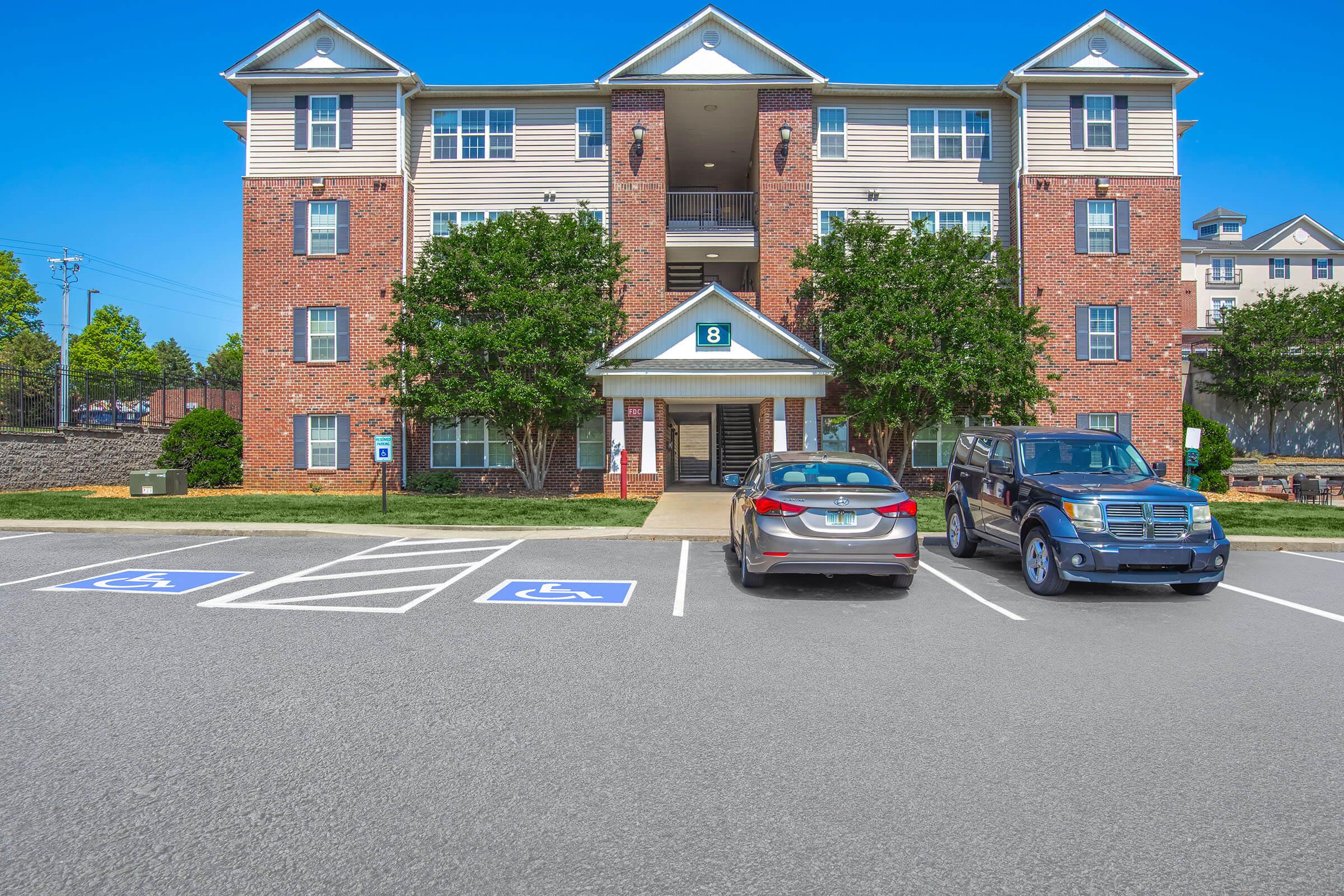 A three-story residential building with a brick facade and light-colored siding. The entrance is flanked by two trees, and there are multiple accessible parking spaces marked out front. Two cars are parked in the lot, one gray and one black. Clear blue sky in the background.