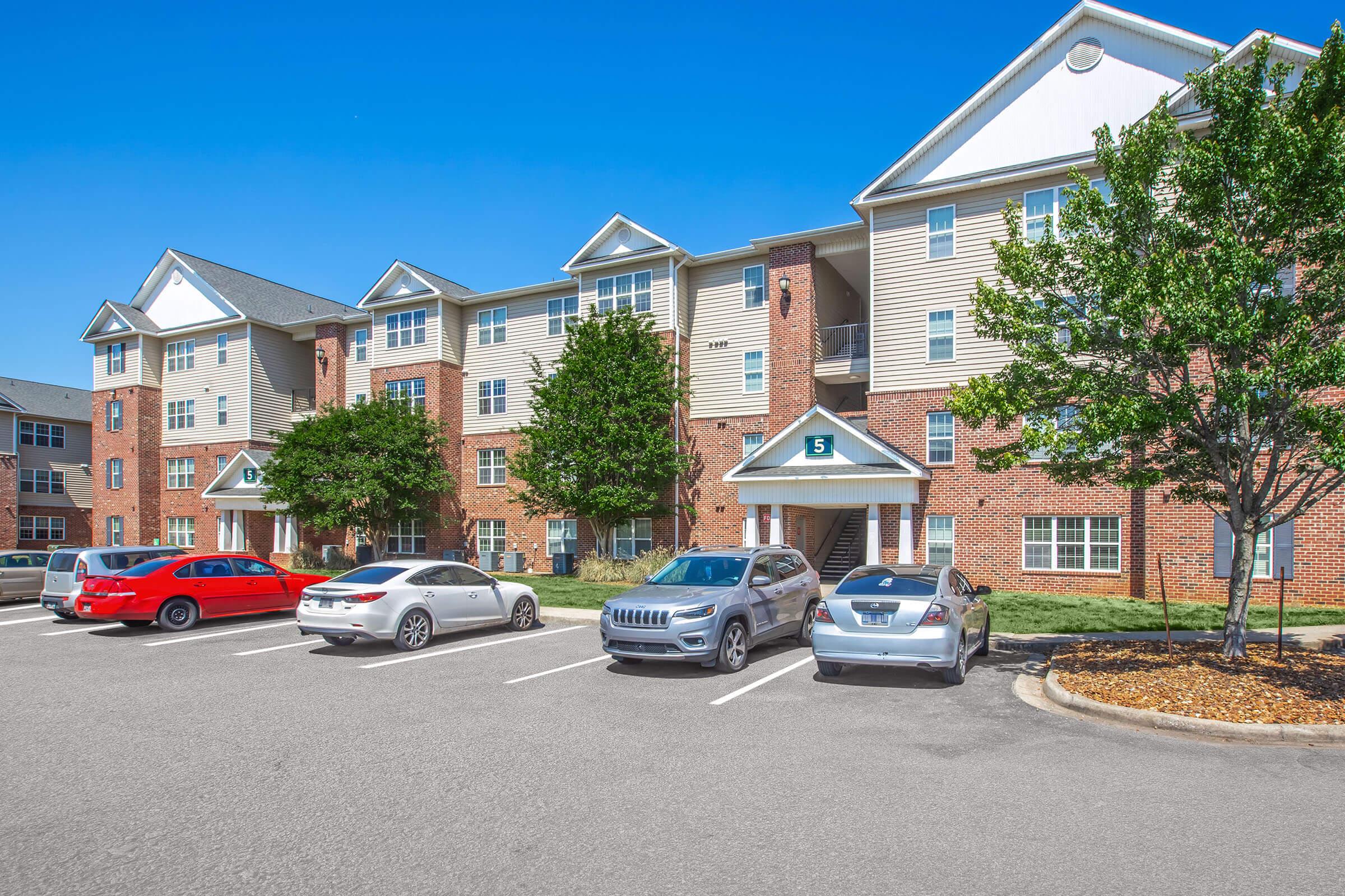A modern three-story apartment building with brick exterior and multiple windows. The front features green trees and a parking lot with several cars parked, including red, silver, and gray vehicles. The sky is clear and blue, indicating a sunny day.