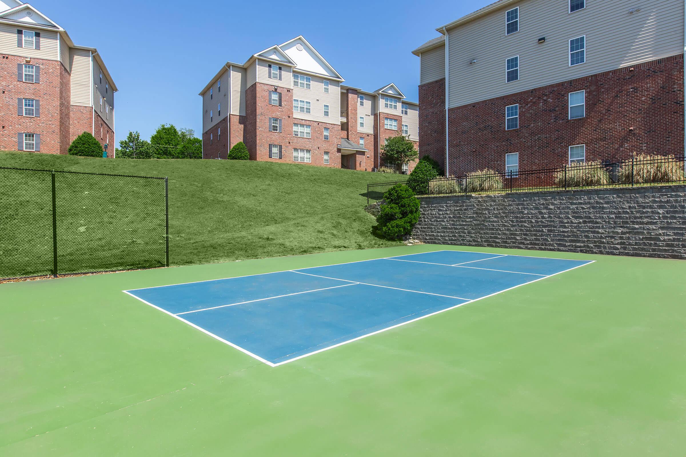 A view of a tennis court with blue markings on a green surface, situated in an outdoor area. In the background, there are multi-story brick apartment buildings and a grassy hill. The sky is clear and blue, creating a bright and open atmosphere.