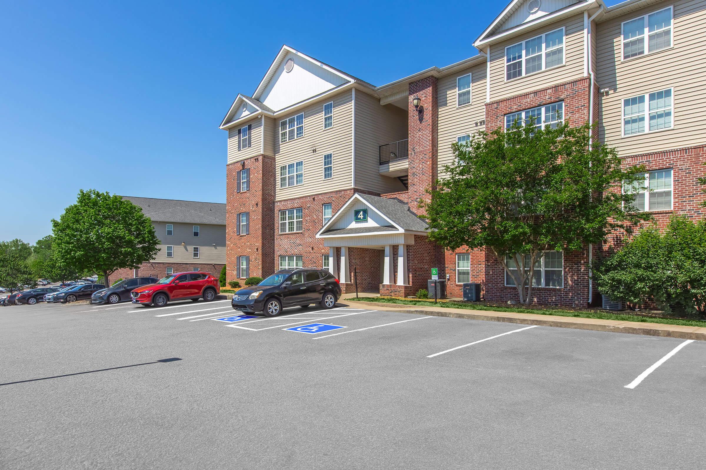Exterior view of a modern apartment building with a brick facade. The building features multiple stories, several windows, and a canopy entrance. A parking lot is visible in the foreground with designated accessible parking spaces. Clear blue sky is in the background, enhancing the bright and inviting atmosphere.