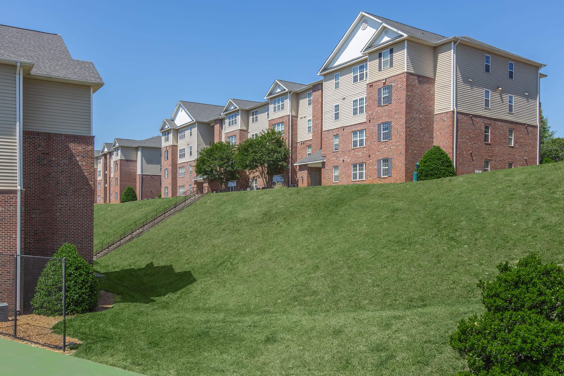 A row of multi-unit residential buildings made of brick and siding, situated on a green, grassy slope under a clear blue sky. The structures feature multiple windows and balconies, with neatly trimmed shrubs in the foreground. Stairs lead up the hillside between the buildings.