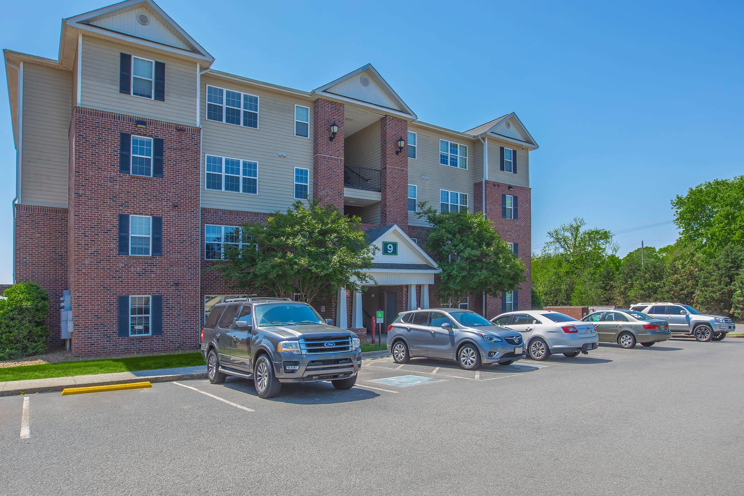 A multi-story brick apartment building with a light-colored façade, featuring windows on multiple levels. There are several parked vehicles in front, including SUVs and sedans. Green trees line the side, and the sky is clear and blue.