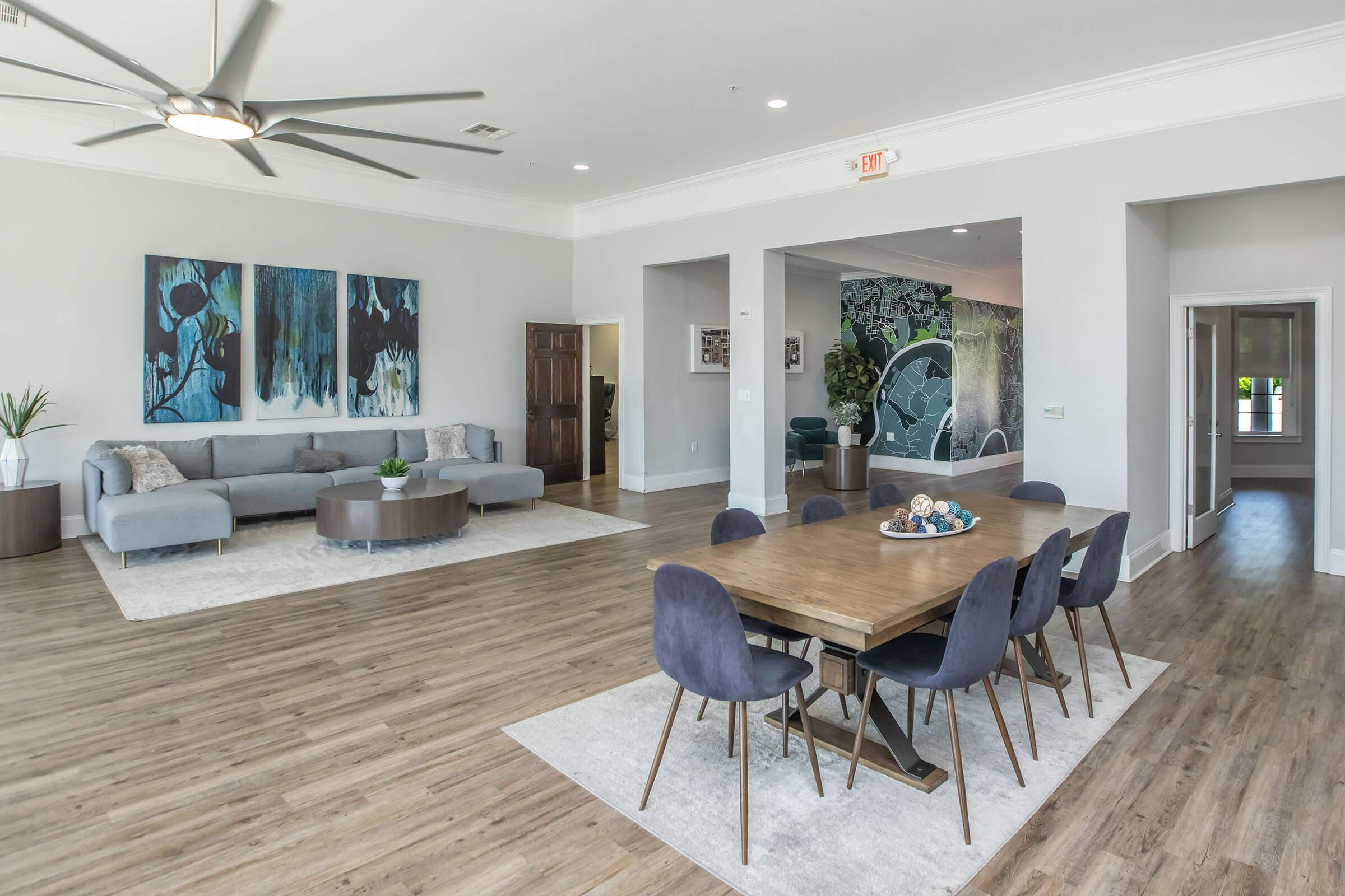 Spacious modern living area featuring a light gray sectional sofa, a round coffee table, and a dining table with six chairs. The walls are adorned with colorful artwork, and large windows provide natural light. The flooring is light wood, enhancing the contemporary design.