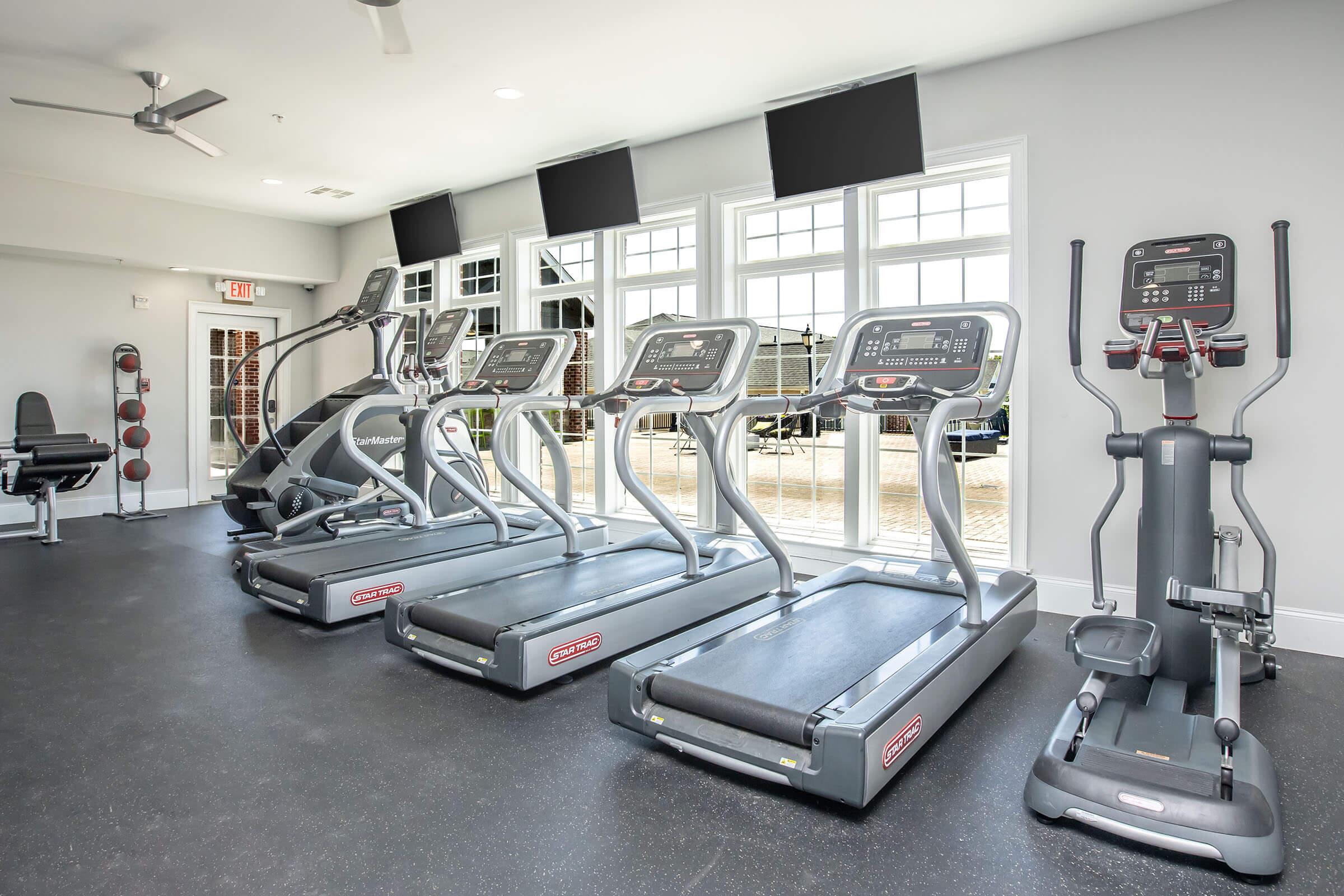 A modern gym interior featuring multiple treadmills, an elliptical machine, and fitness equipment. The space has large windows allowing natural light, with mounted TVs on the walls and a clean, minimalist design. The flooring is dark, and there are additional exercise items in the background.