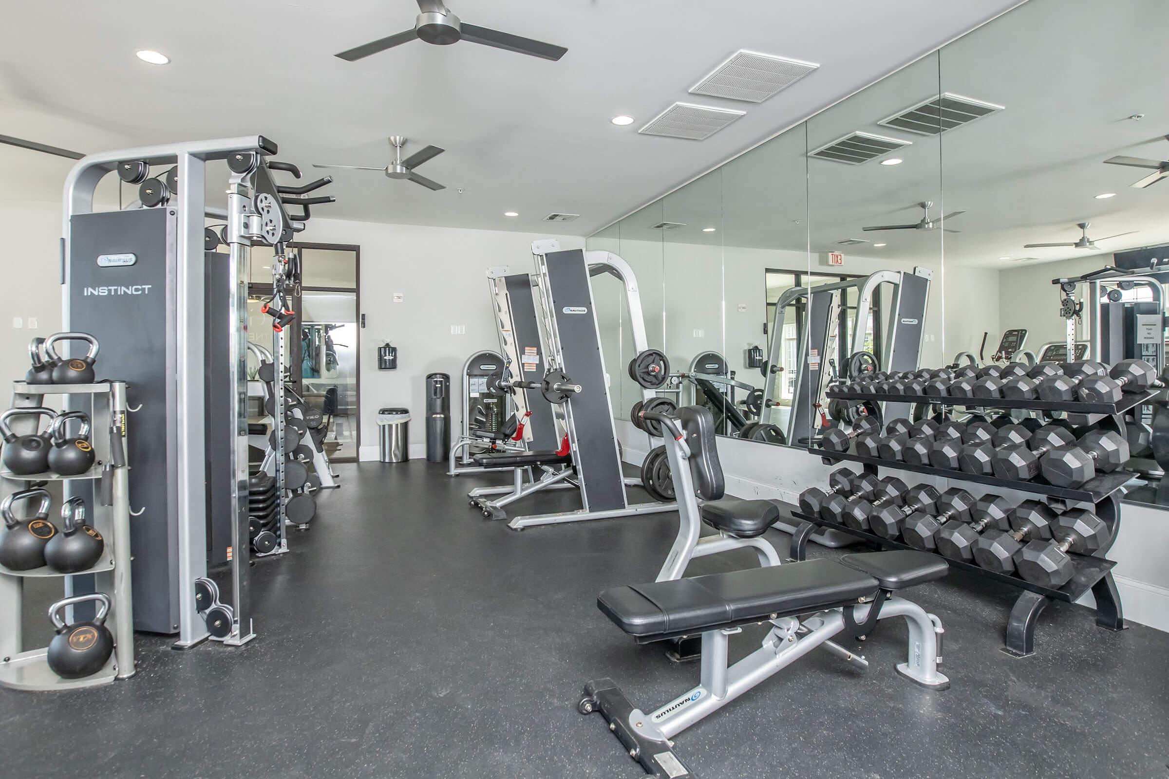 A modern gym interior featuring various weight training equipment, including dumbbells arranged on a rack, a weight bench, cable machines, and a mirror wall, with fans for ventilation. The space is well-lit and designed for fitness workouts.
