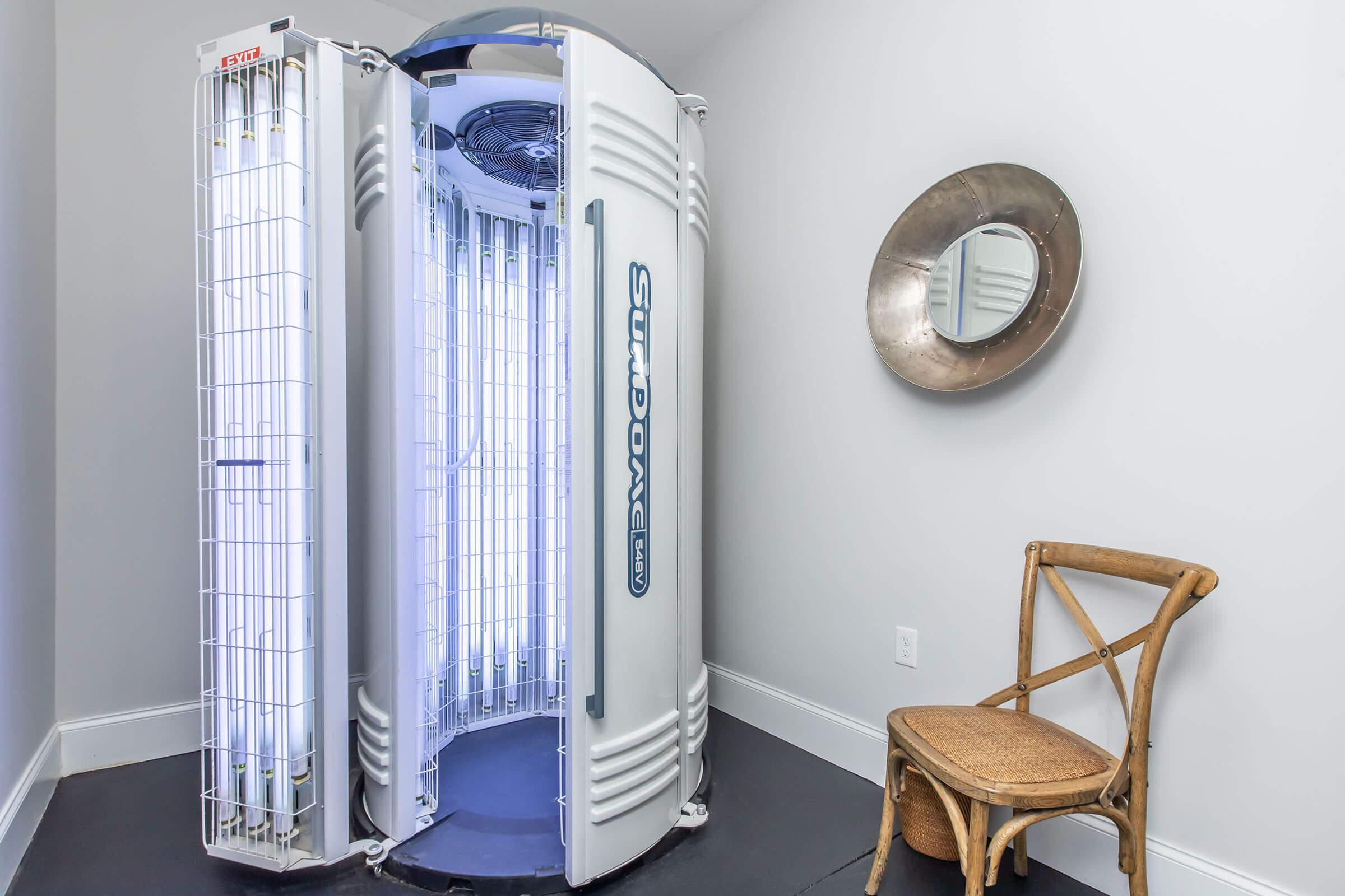 A sun booth with bright fluorescent lights for tanning, positioned in a room with neutral-colored walls. Next to it, there is a modern round mirror and a wooden chair with a simple design. The environment appears clean and well-lit.