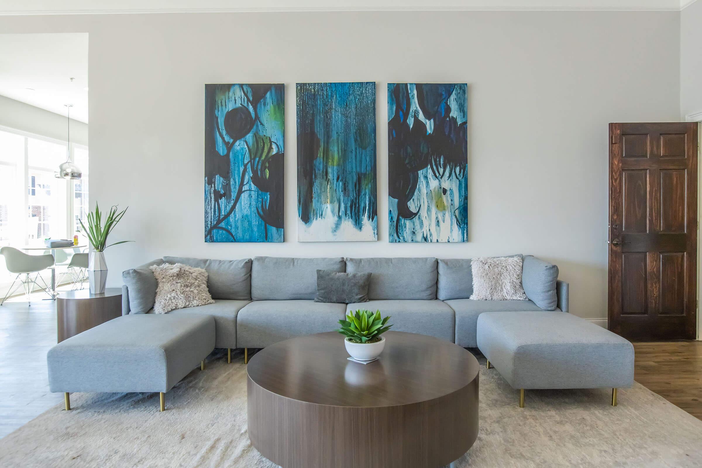 A stylish living room featuring a gray sectional sofa with decorative pillows, a round wooden coffee table, and a small plant centerpiece. The walls are adorned with three large abstract paintings in blue and black tones. Natural light fills the space, enhancing the modern decor. A wooden door is visible on the right.