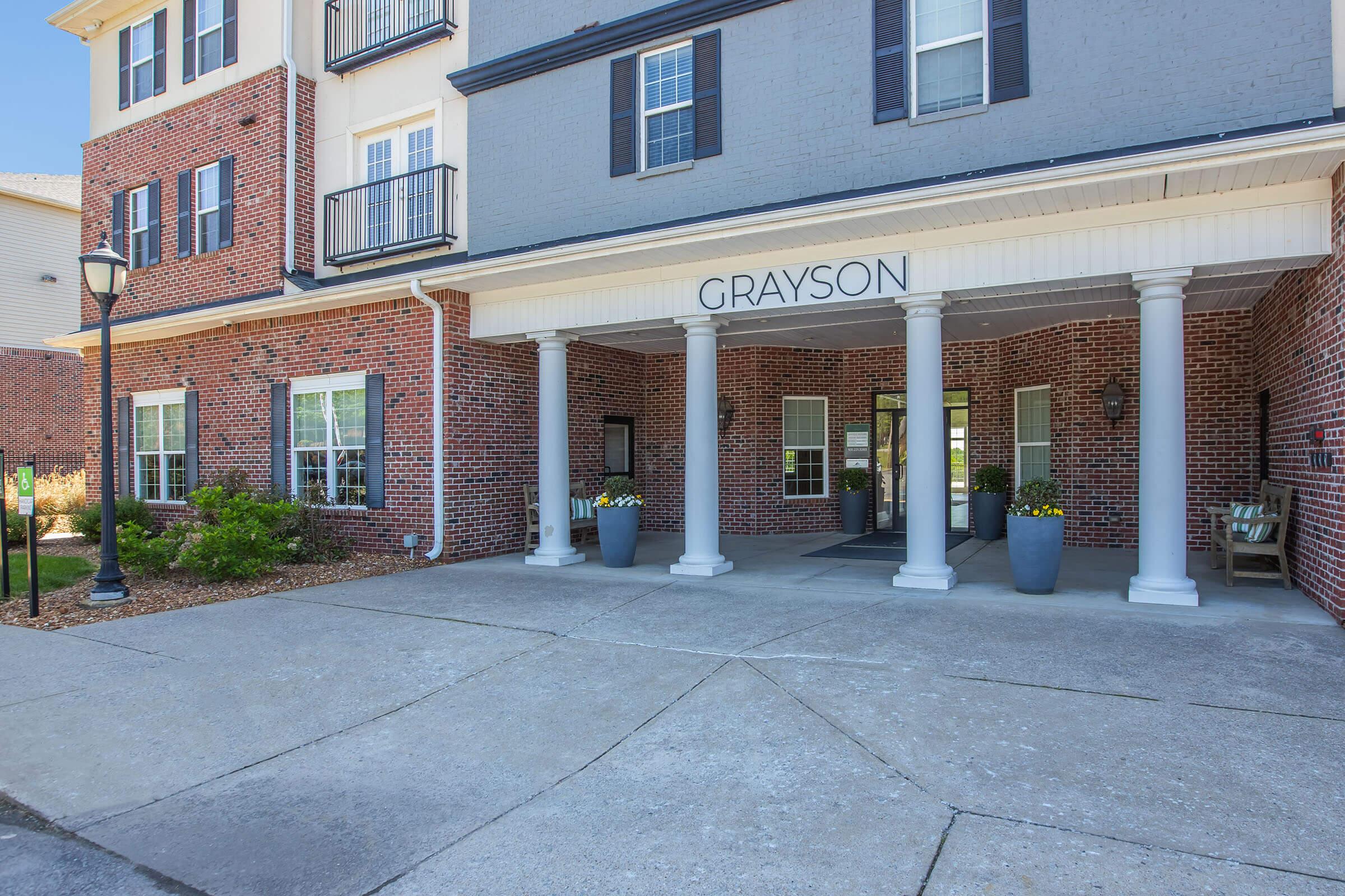 Entrance of a building named "GRAYSON," featuring a covered porch with white columns. The façade includes a mixture of brick and painted siding, with large windows on either side. Potted flowers decorate the entrance area, and there are outdoor light fixtures lining the walkway.