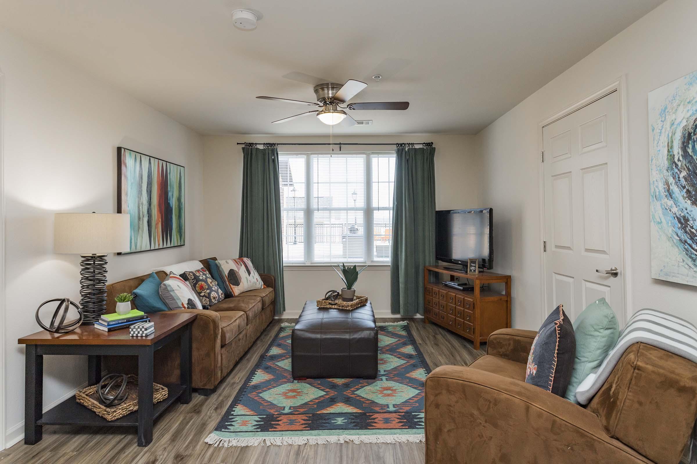 A cozy living room featuring a brown sofa with colorful throw pillows, a dark coffee table, and a decorative rug. There's a lamp on a side table, a green plant, and a television on a stand. Large windows with light green curtains allow natural light to fill the space, complemented by wall art in vibrant colors.