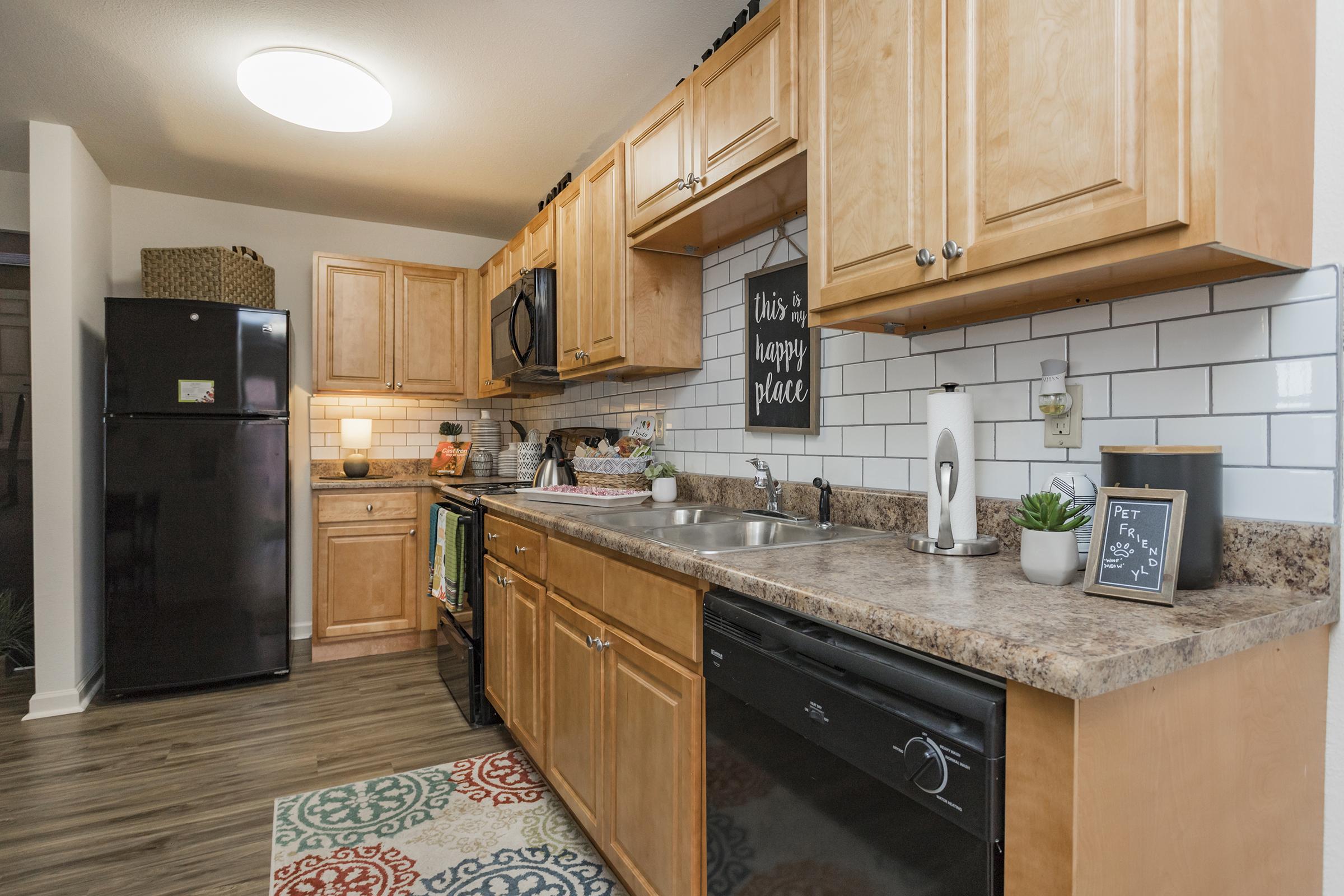 A modern kitchen featuring wooden cabinets, a black refrigerator, a stainless steel sink, and a dishwasher. The backsplash has white subway tiles, and there's a decorative sign on the wall. A rug with a colorful design is on the floor, and various kitchen items and plants are on the countertop.