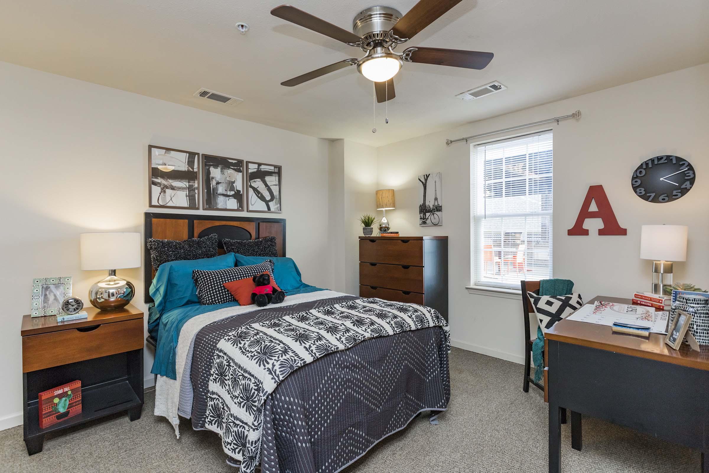 A cozy bedroom featuring a queen-sized bed with decorative pillows and a patterned blanket, a modern dresser, a desk with a chair, and a window with natural light. Wall art and a clock add character, and there’s a ceiling fan for ventilation. The decor is stylish and inviting.