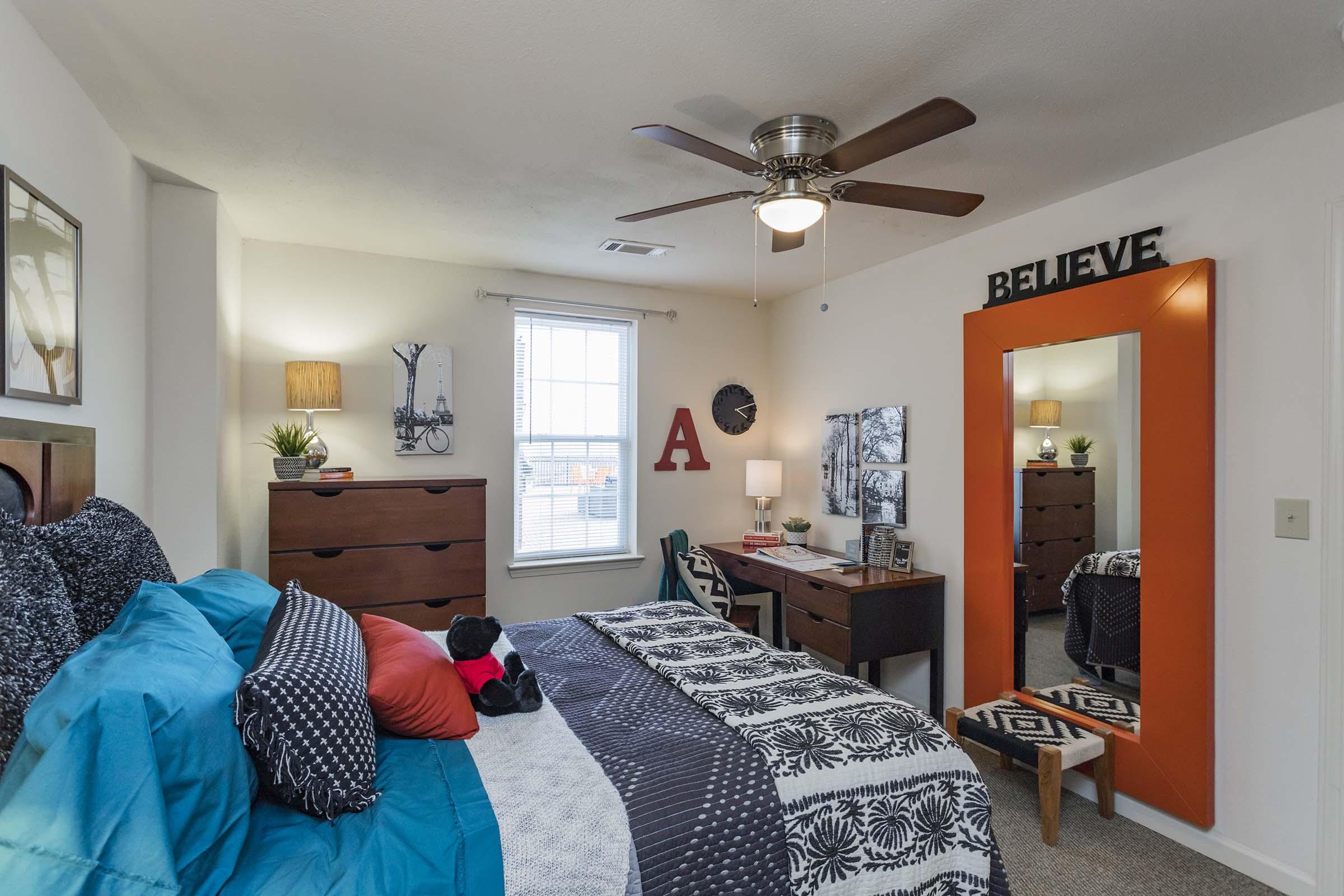 A cozy bedroom featuring a queen-sized bed with blue and black bedding, a wooden dresser, and a desk with a mirror. The room is decorated with a wall clock, plants, and the word "BELIEVE" in black letters on the wall. Natural light enters through a window, creating a warm atmosphere.