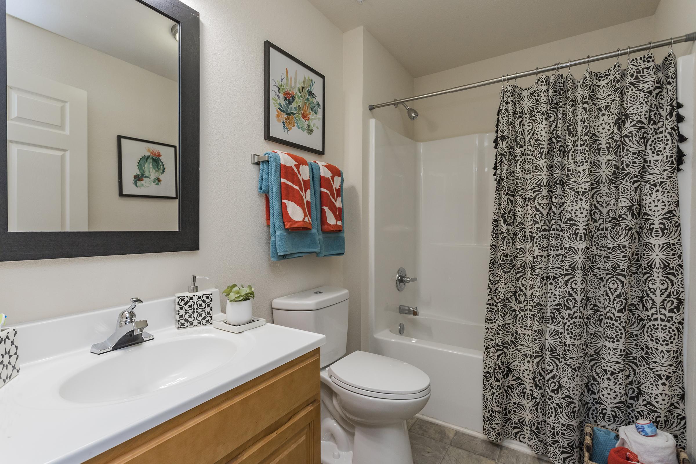 A clean, modern bathroom featuring a white bathtub with shower, a light wood vanity with a sink, and a mirror above it. Decorative wall art is displayed, along with vibrant towels hanging on a towel rack, and a patterned shower curtain. Plants and toiletries are neatly arranged on the countertop.