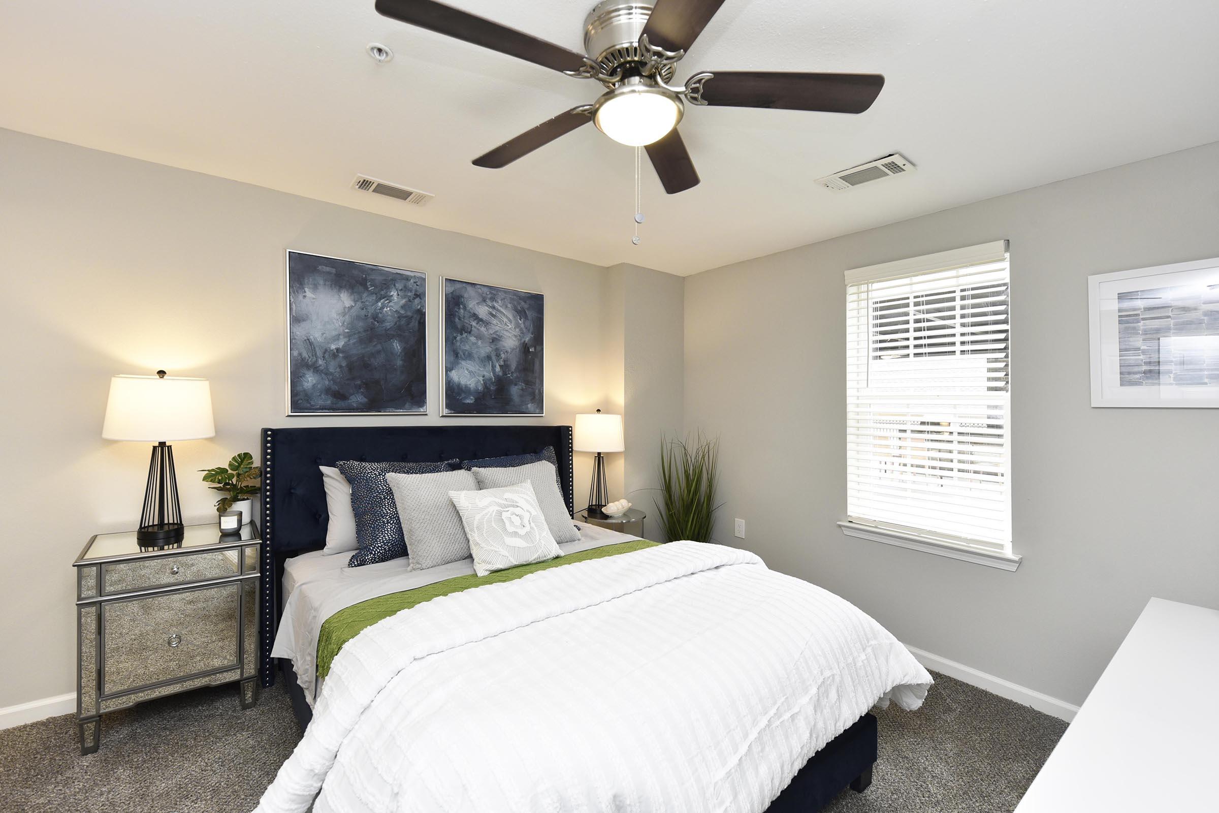 A cozy bedroom featuring a navy blue upholstered bed with white bedding, two decorative pillows, and a green throw blanket. There's a stylish mirrored nightstand with a lamp on one side, and on the other side, a window with white blinds. Artwork is hung above the bed, adding a contemporary touch to the decor.