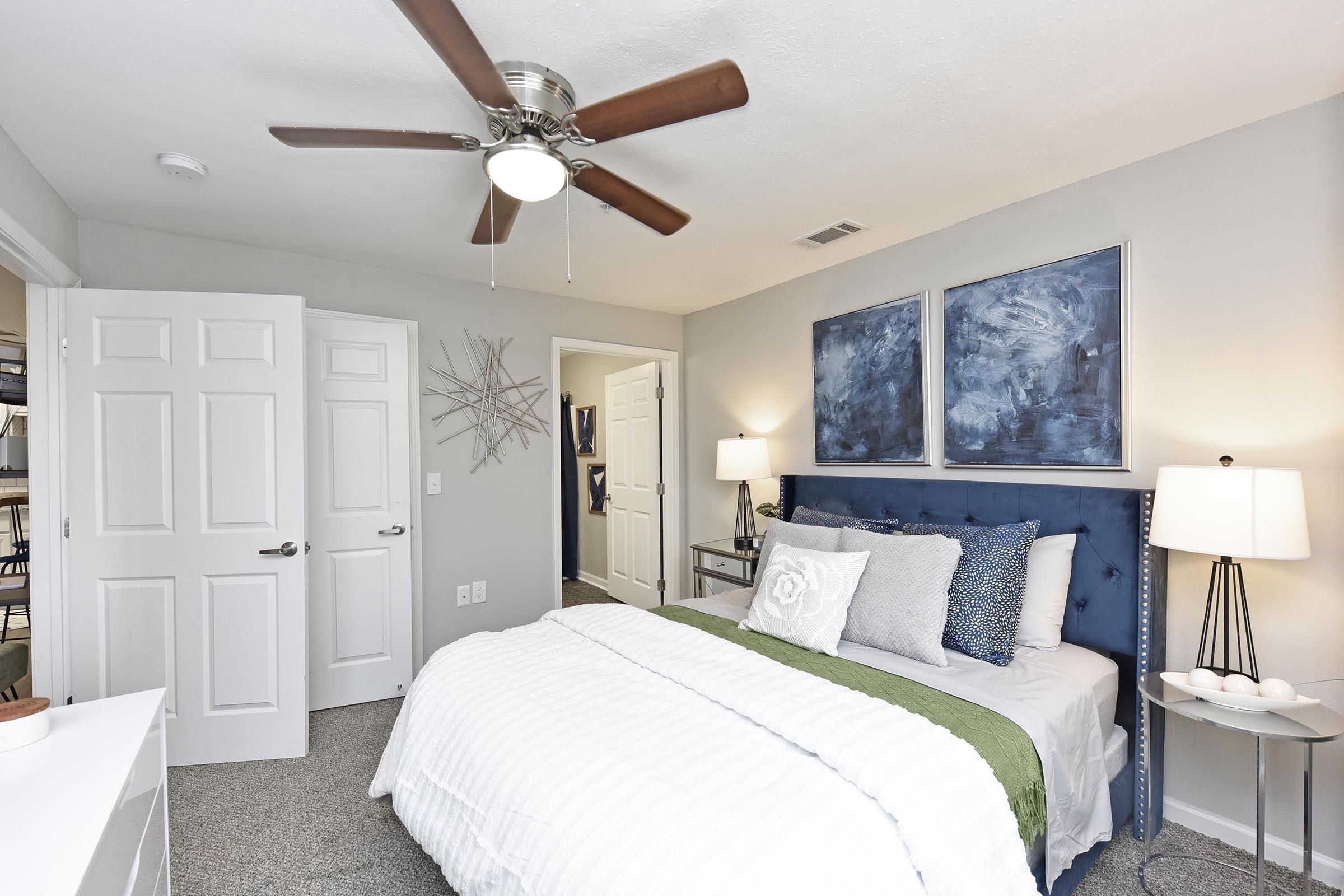 A cozy bedroom featuring a white bedspread on a neatly made bed with decorative pillows. There are blue abstract paintings on the wall above the bed, and a bedside table with a lamp on each side. A ceiling fan hangs above, and a closet is visible in the background through an open door.