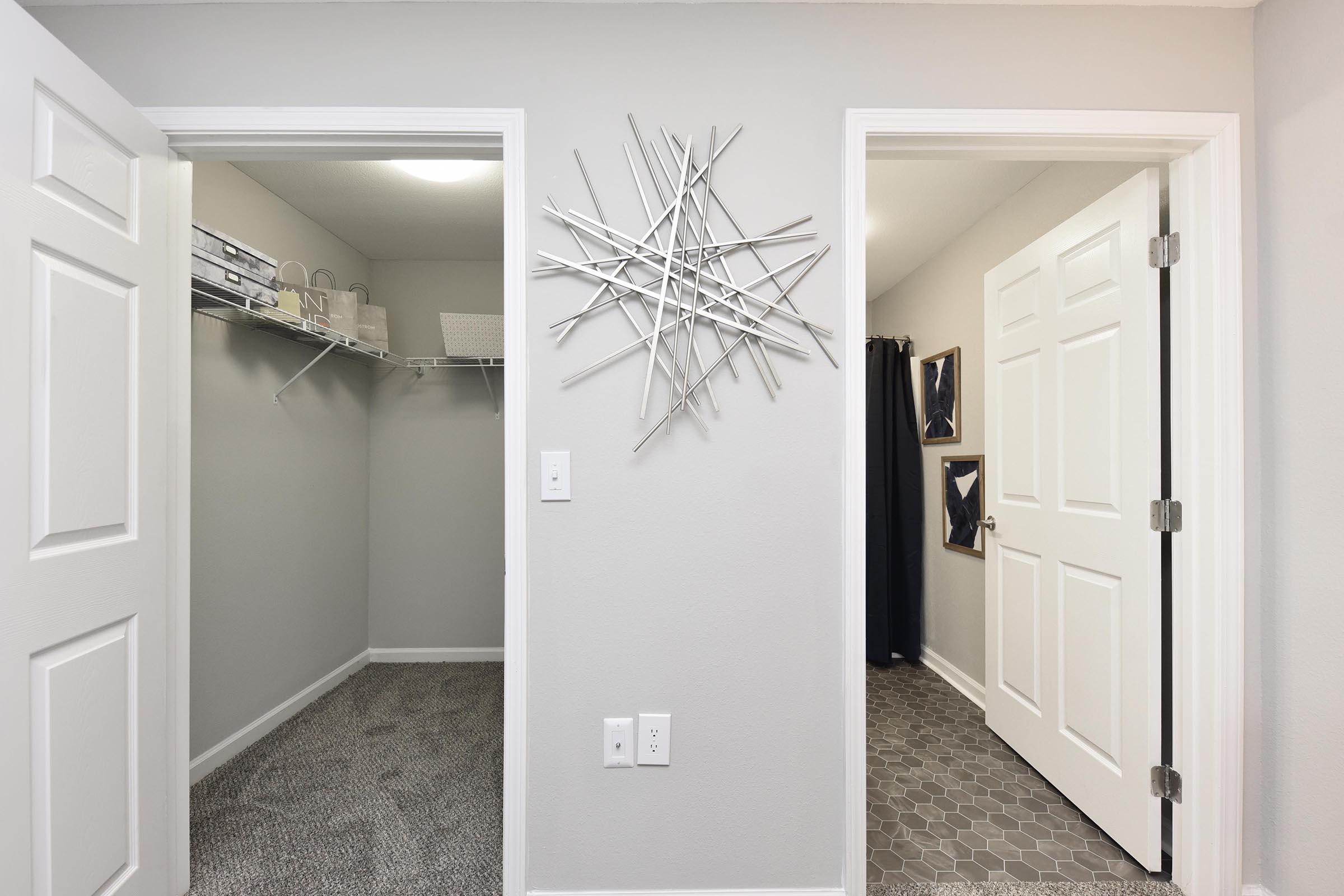 A well-lit hallway leading to two open doors: one door opens to a spacious walk-in closet with shelves, while the other leads to a room featuring a geometric wall decoration and framed artwork. The walls are painted gray, and the flooring is a combination of carpet and patterned tiles.