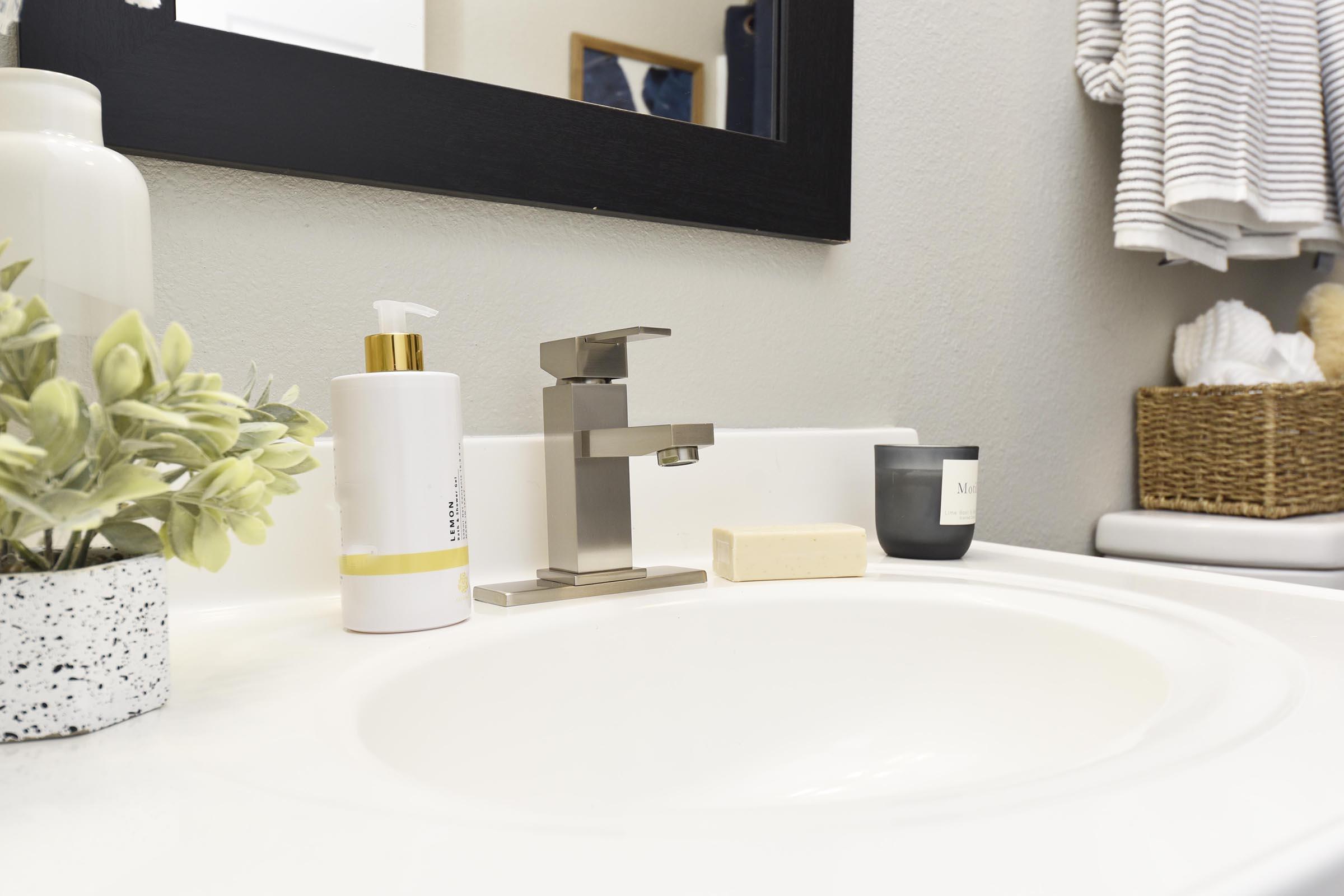 A modern bathroom sink with a sleek faucet, a bottle of liquid soap, a bar of soap, a small candle, and a potted plant. In the background, there are neatly arranged towels and a woven basket. The walls are painted a light color, creating a clean and contemporary aesthetic.