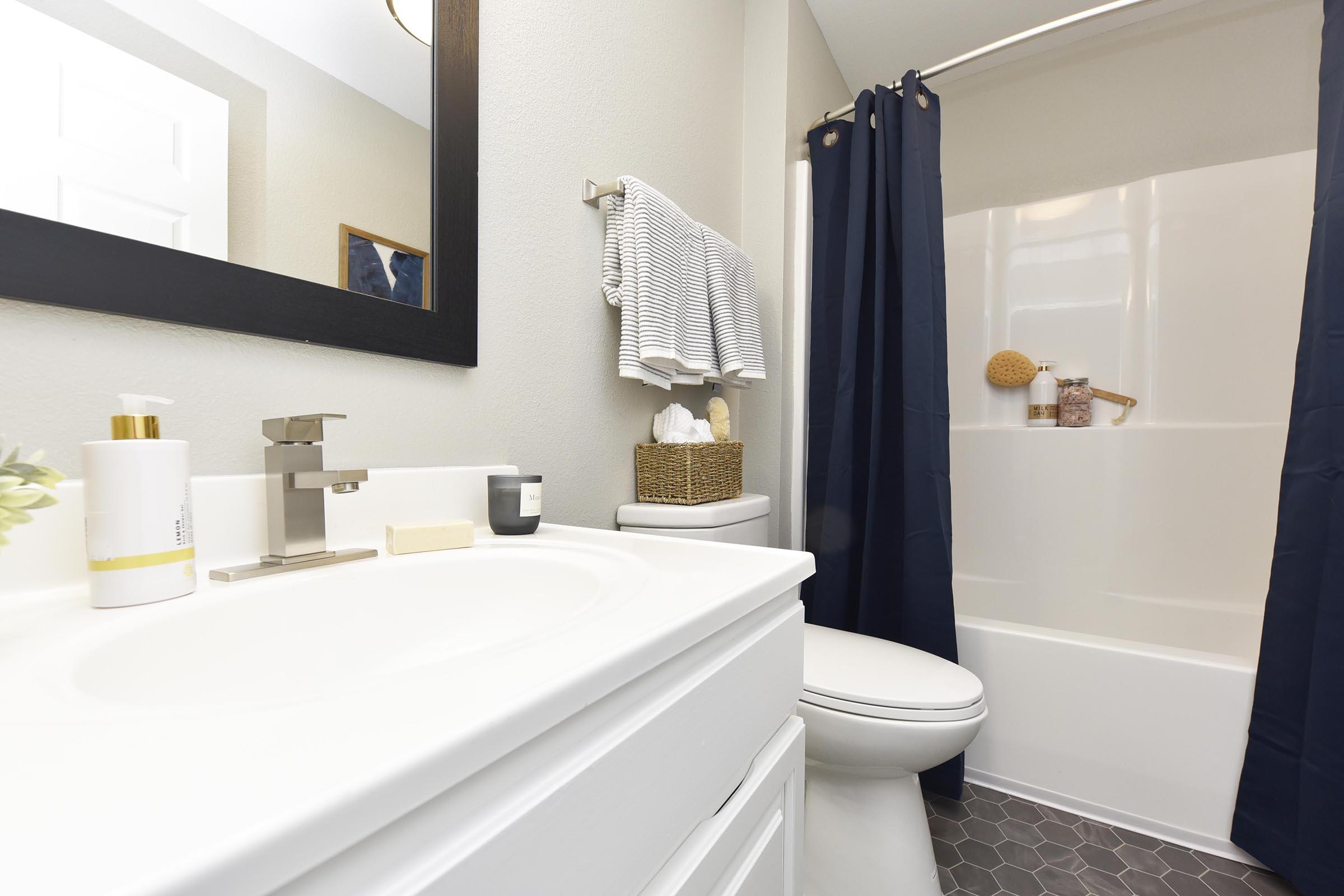 A clean bathroom featuring a white countertop with toiletries, a modern sink faucet, a mirror with a black frame, and a shower area with a white curtain. Decorative items include a woven basket, towels, and a small plant on the counter, creating a minimalist and welcoming atmosphere.