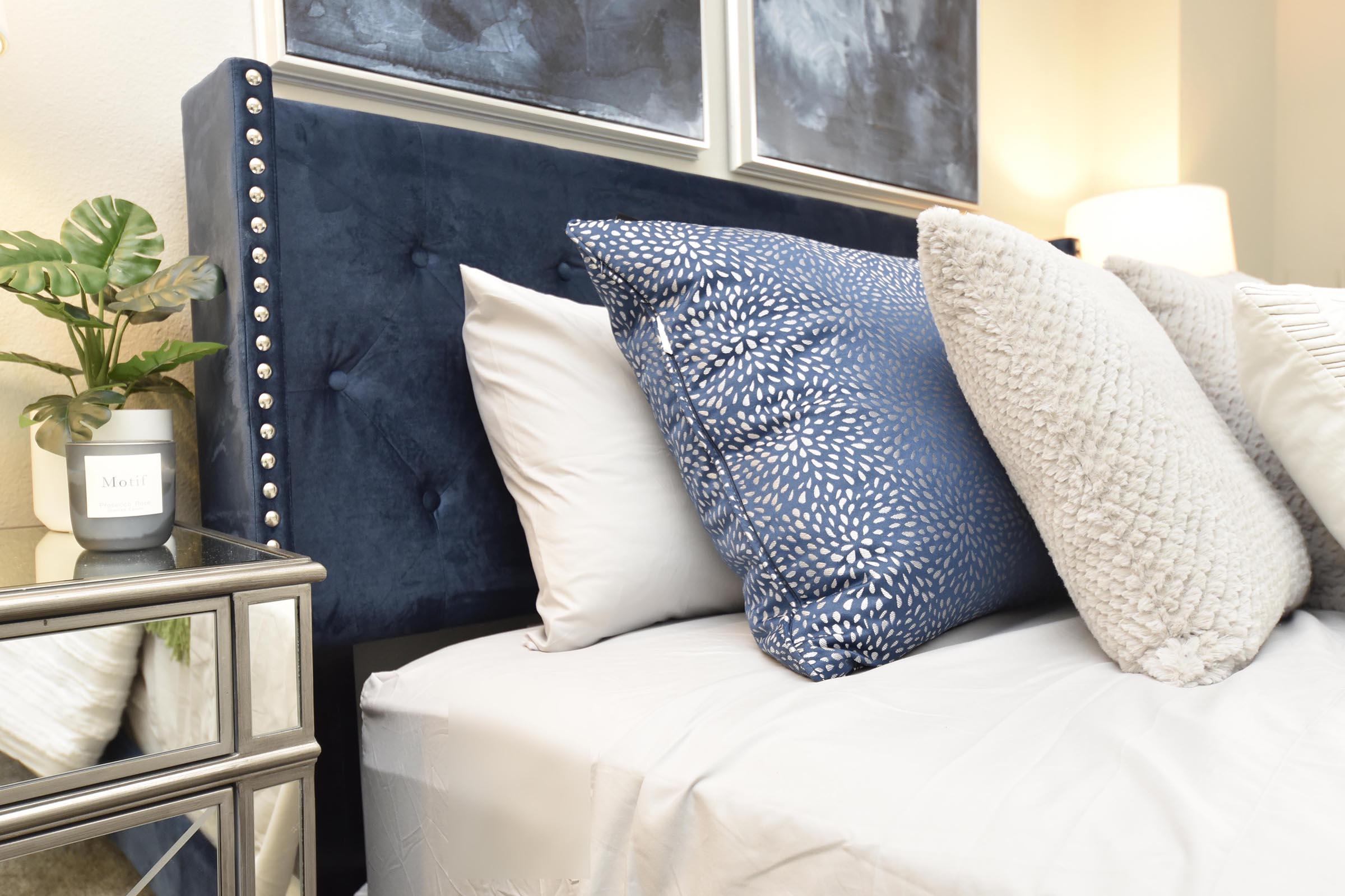 A cozy bedroom scene featuring a plush bed with an upholstered navy blue headboard adorned with silver studs. The bed is layered with multiple decorative pillows in varying textures and colors. A bedside table with a potted plant and a candle adds to the inviting atmosphere, complemented by artistic wall decor above the bed.