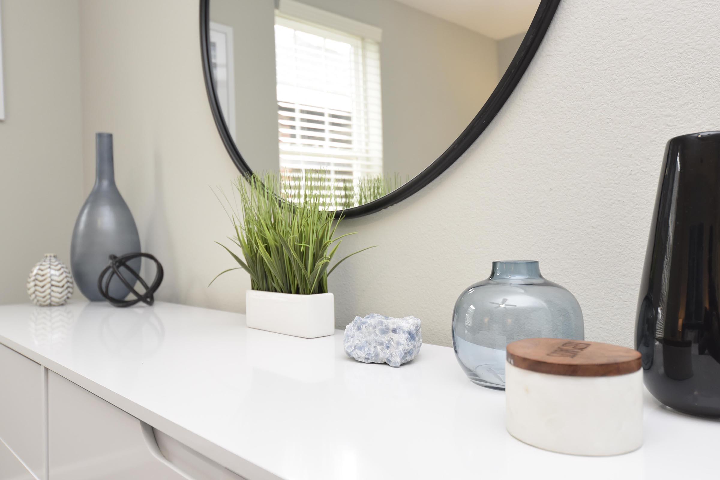 A modern interior scene featuring a white surface with a variety of decorative items: a tall, gray vase, a small textured blue vase, a white planter with green grass, a round wooden container, a blue decorative stone, and a black vase, all displayed near a circular mirror reflecting natural light.
