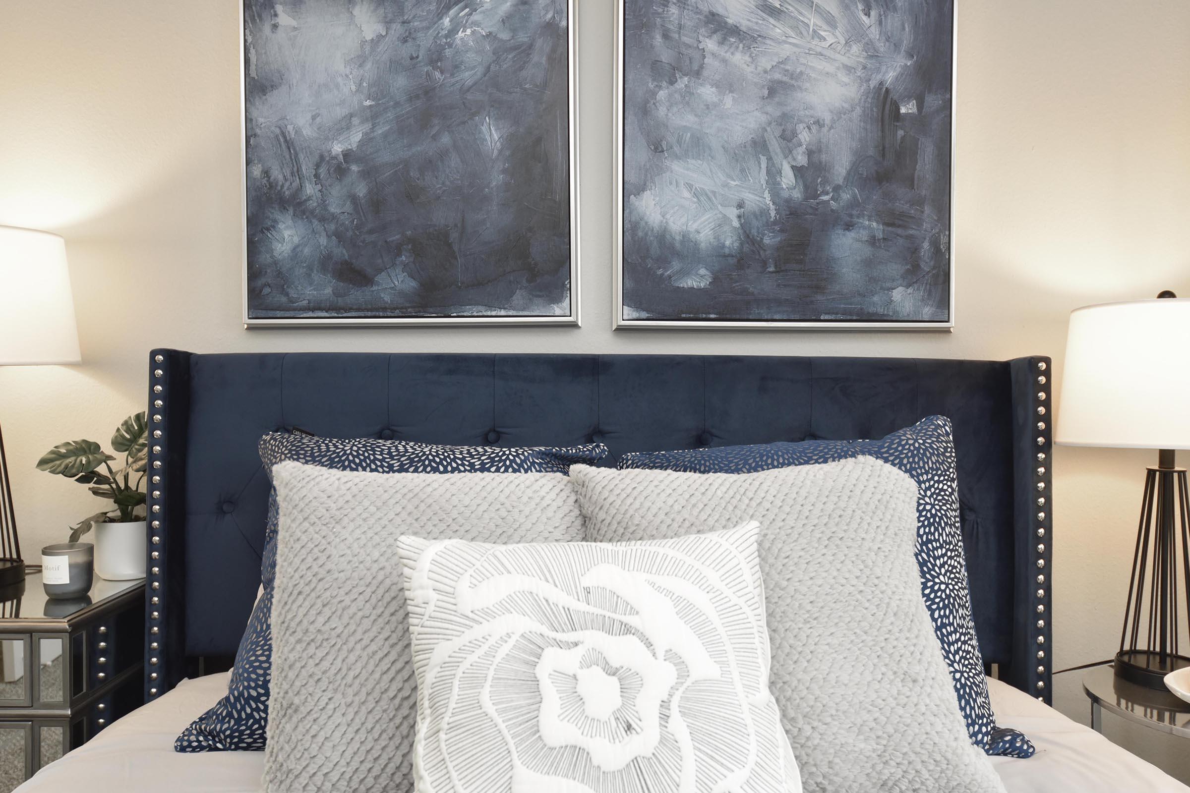 A stylish bedroom featuring a navy blue upholstered headboard with decorative silver studs. The bed is adorned with various textured pillows in shades of gray and blue. Above the headboard are two modern abstract paintings with dark blue hues. On either side of the bed, there are lamps and a small plant on a bedside table.