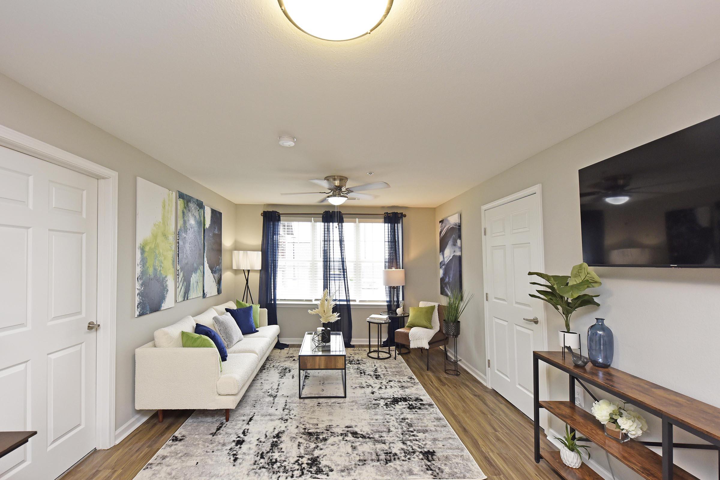 A modern living room featuring a light-colored sofa with decorative pillows, a coffee table, and a stylish area rug. The room has two large windows with blue curtains, artwork on the walls, a ceiling fan, and a small seating area with a chair and potted plant. A flat-screen TV is mounted on the wall.