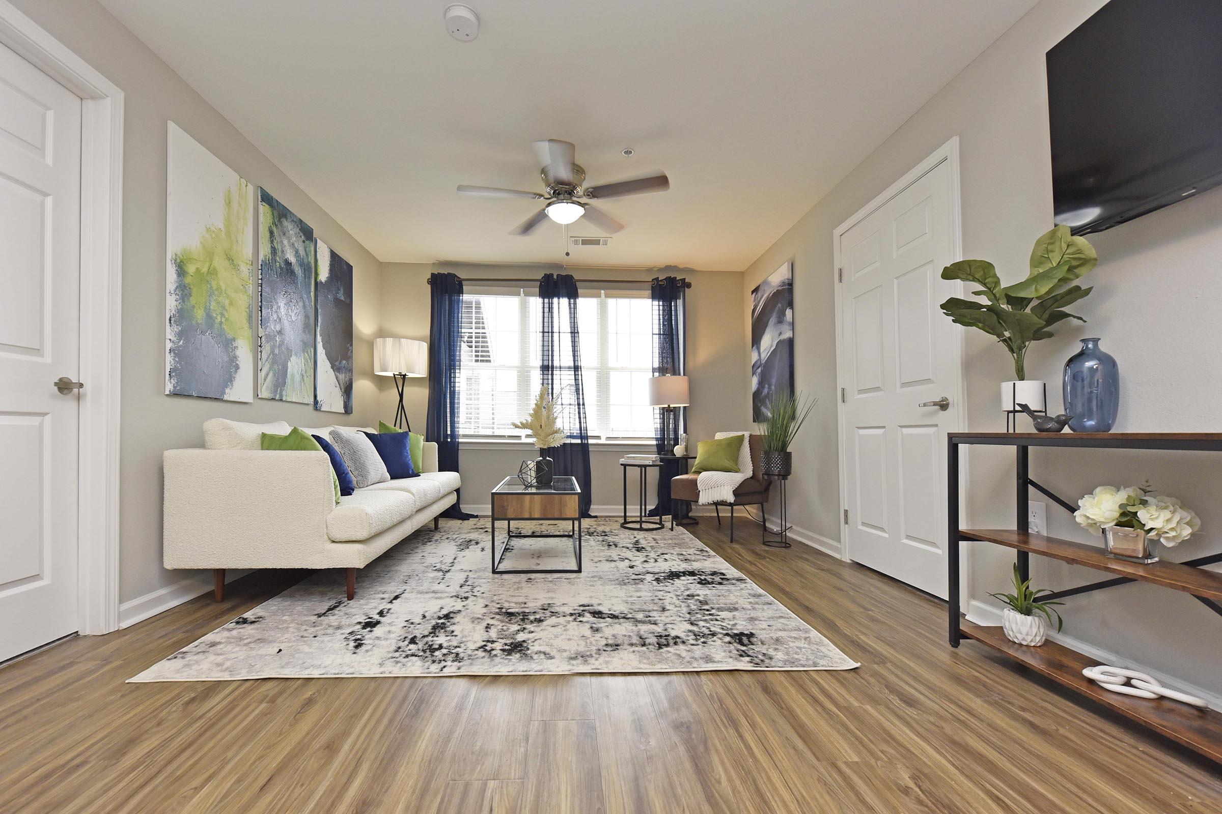 A modern living room featuring a light sofa, decorative pillows, and an area rug. There are large abstract paintings on the walls, a coffee table in the center, and a potted plant in the corner. Natural light floods in through a large window, complemented by curtains. A TV is mounted on the wall.